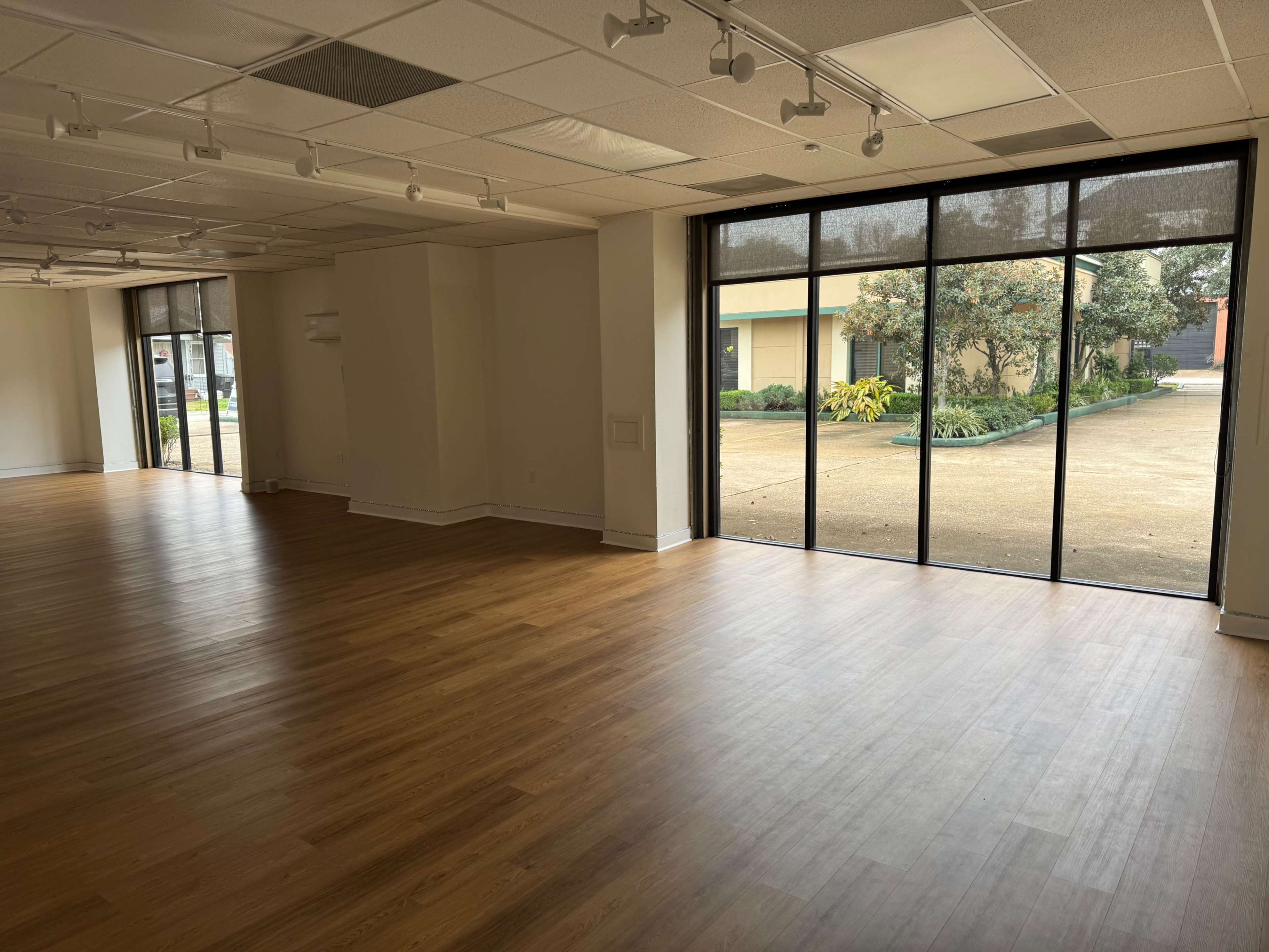 The image shows a spacious, empty room with wooden flooring and large glass doors leading to an outdoor area.