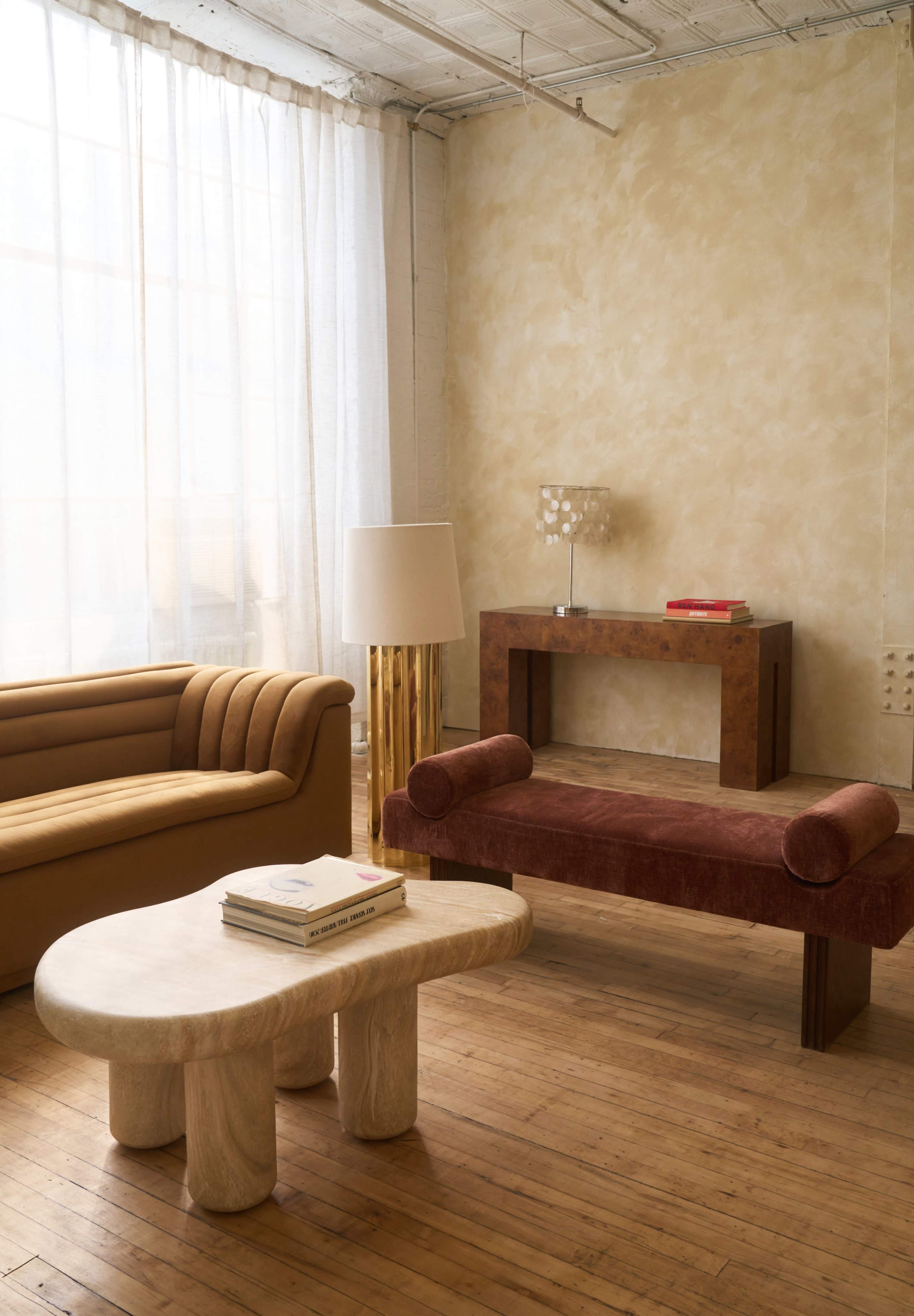 The image shows a minimalist interior featuring a tan sofa, a red upholstered bench, a light wooden coffee table with rounded legs, and a lamp beside a textured wall.