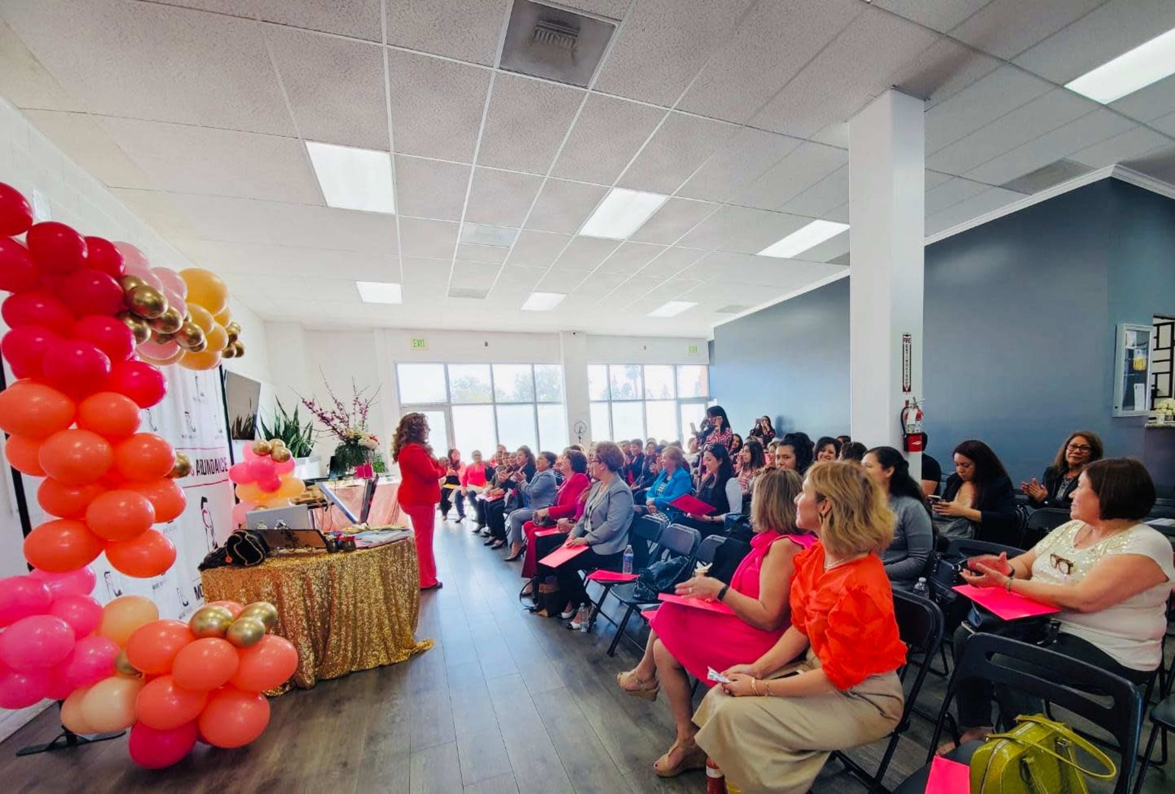 A large group of people is seated in rows facing a speaker in front of a decorated backdrop featuring balloons and a table with a shiny tablecloth.