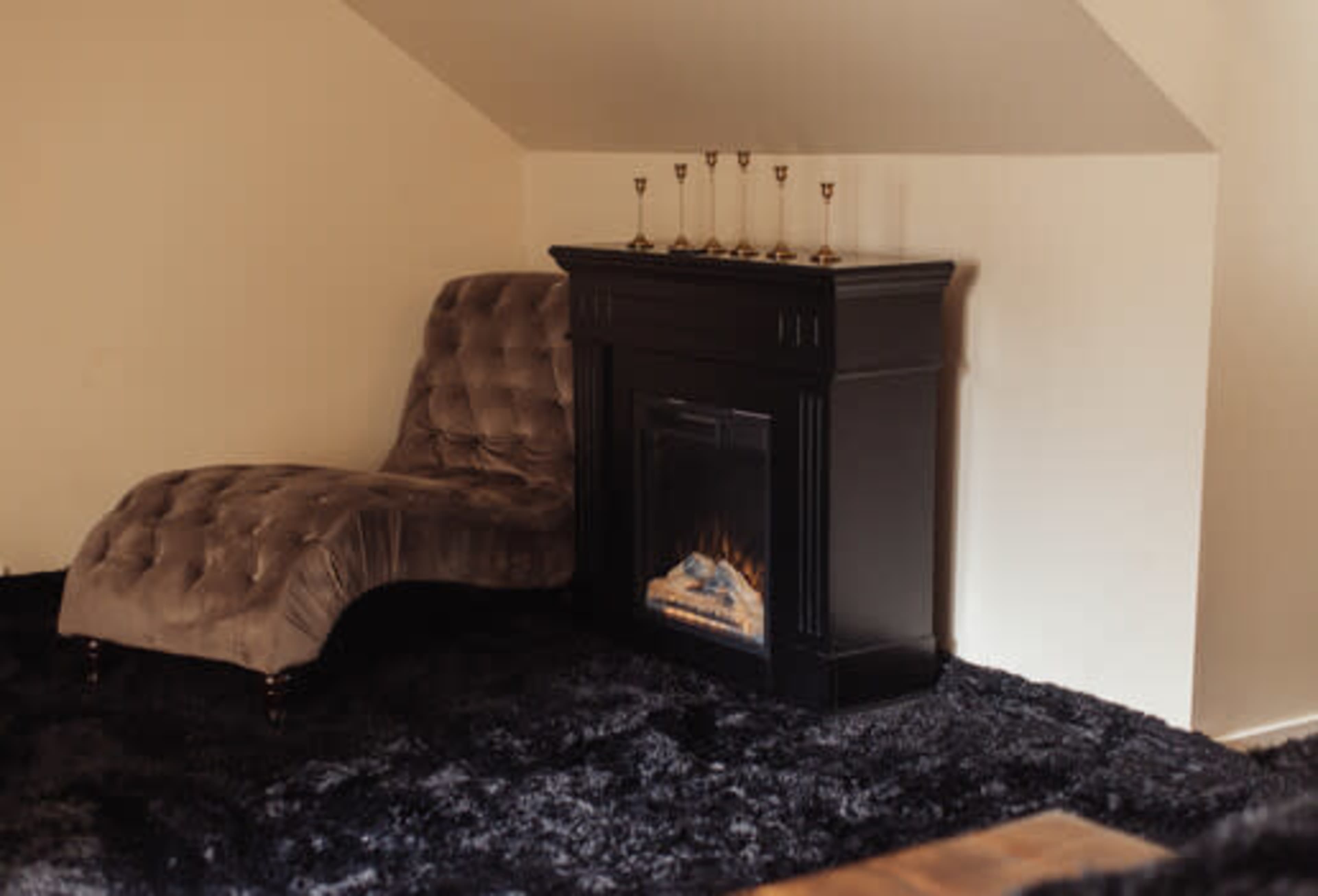 A plush chaise lounge sits next to a black electric fireplace on a dark, shaggy rug in a softly lit room.