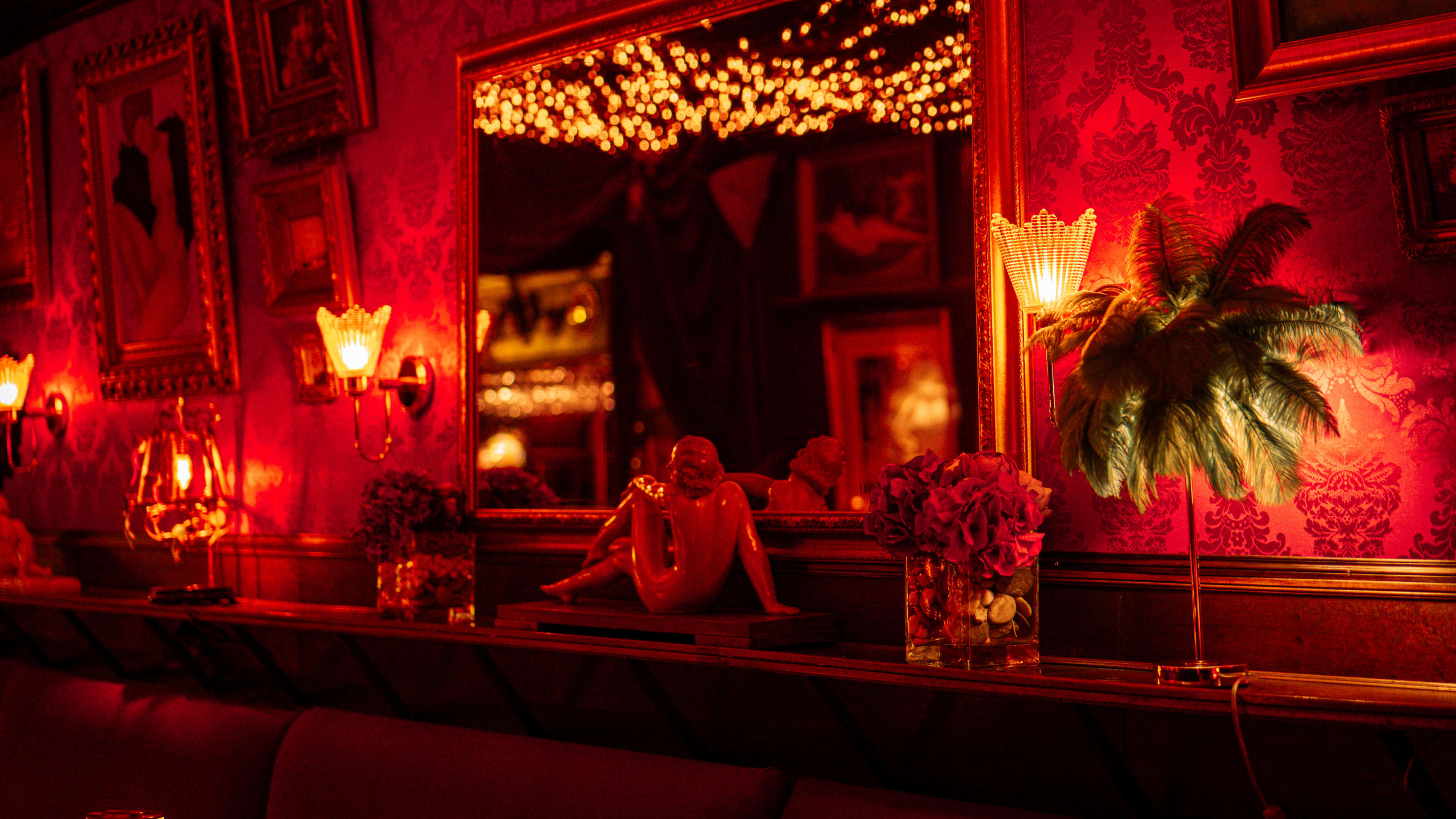 The image features a warmly lit interior of a restaurant or bar, with ornate red wallpaper, decorative mirrors, statuettes, and floral arrangements on a shelf.