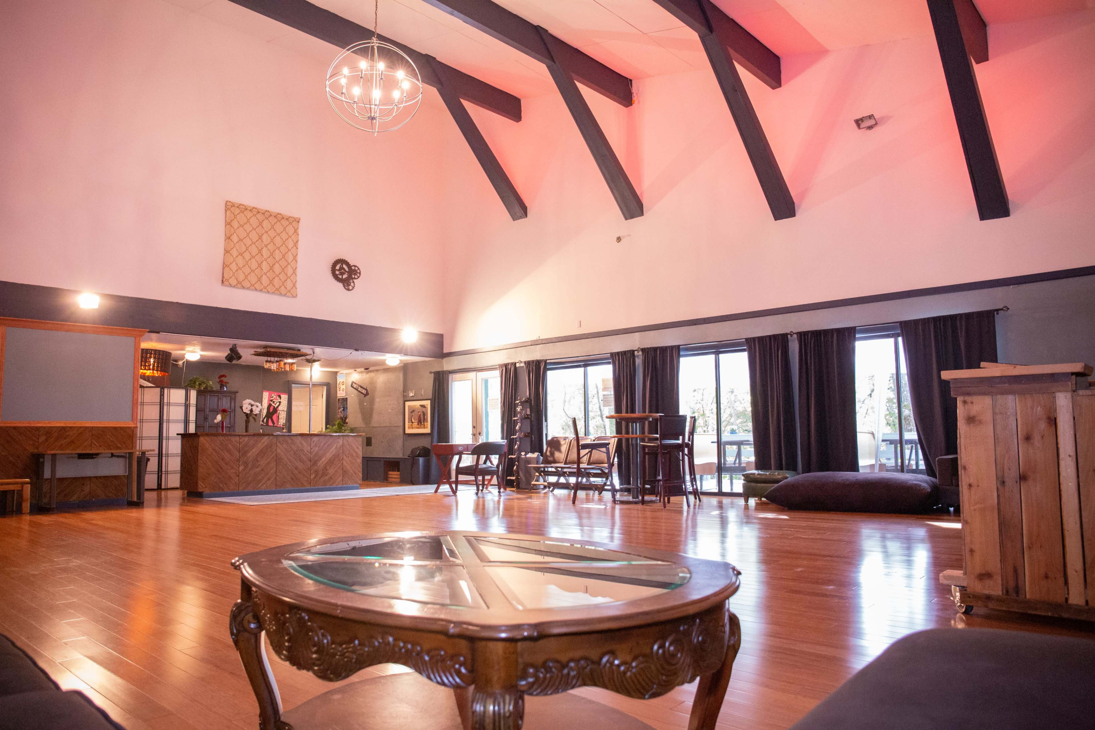 A spacious room with high ceilings features wooden flooring, large windows, and various seating arrangements.
