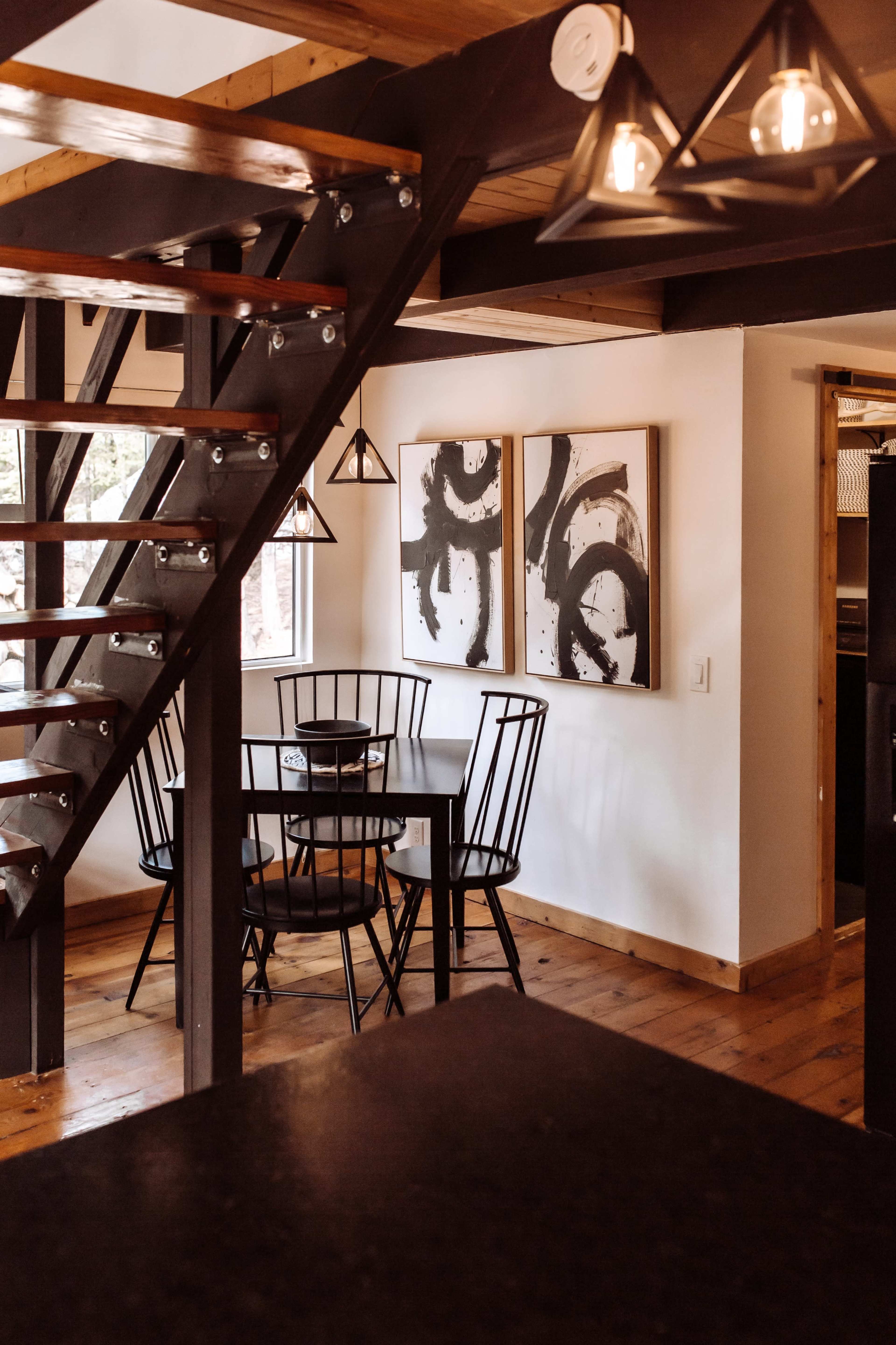 The image shows a dining area with a black table and chairs beneath abstract wall art, adjacent to a staircase with wooden steps and metal railing.