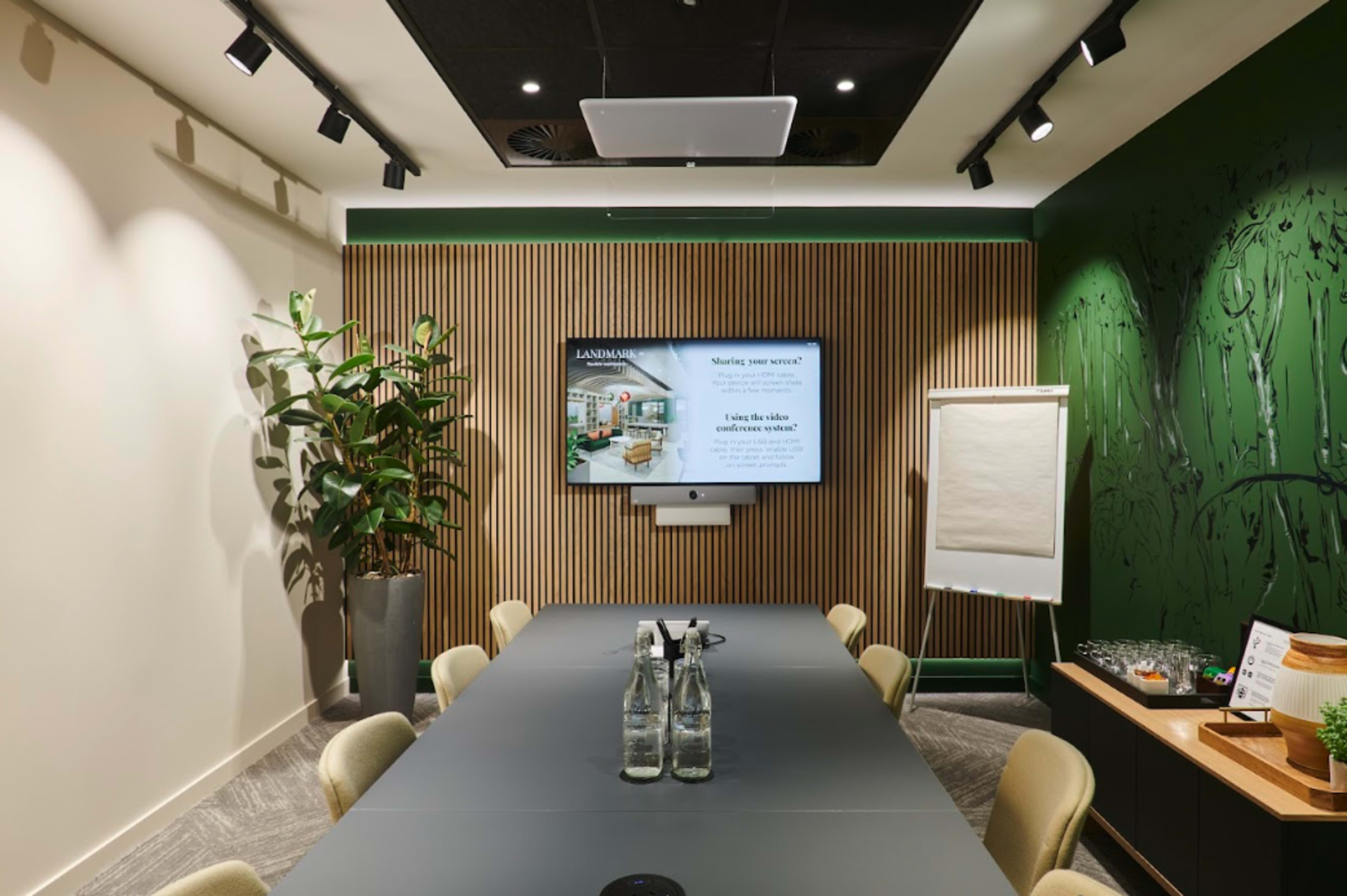 A modern meeting room features a long table, a large screen displaying a presentation, and plants against a wood-paneled wall.