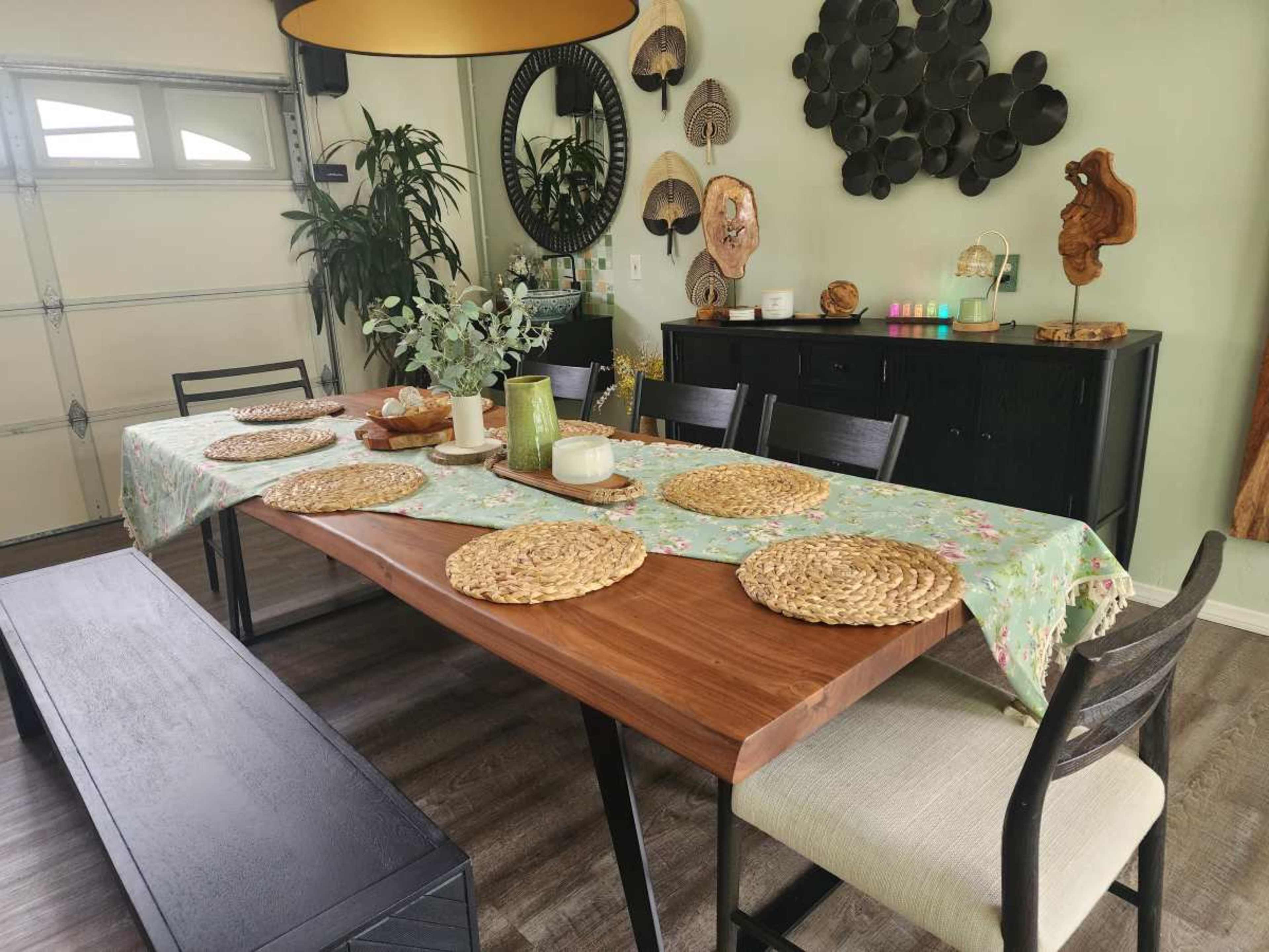 A dining table is set with placemats, cutlery, and a centerpiece in a well-lit room that includes plants and decorative wall art.