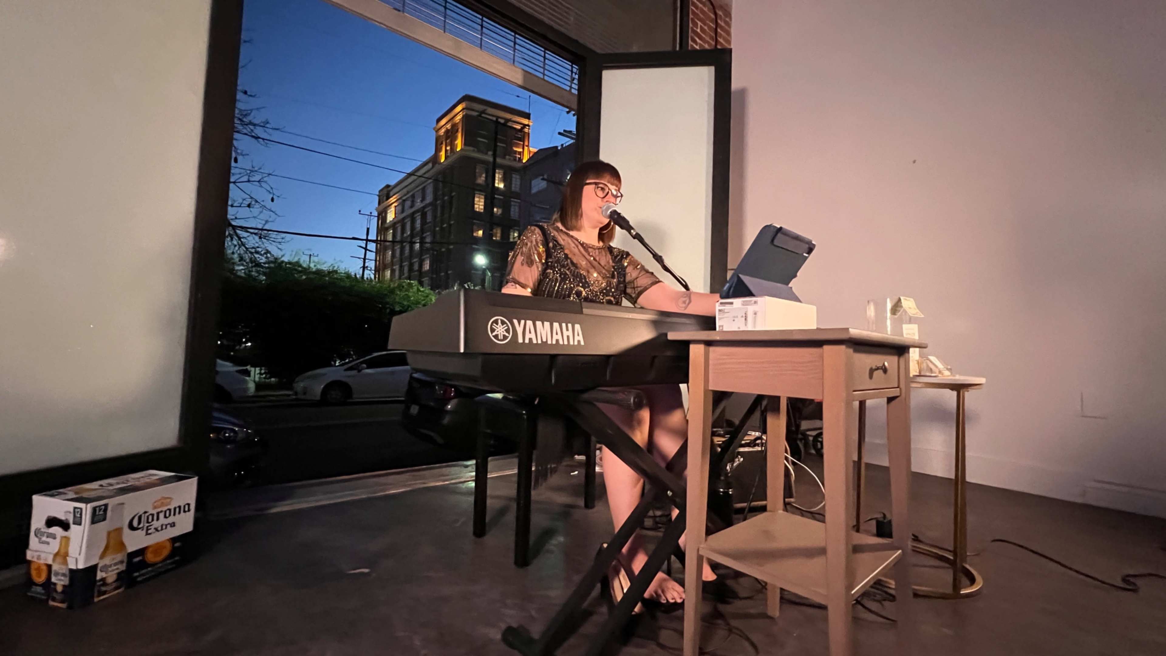A musician performs at a keyboard in a well-lit interior space, with windows showing a twilight cityscape outside.