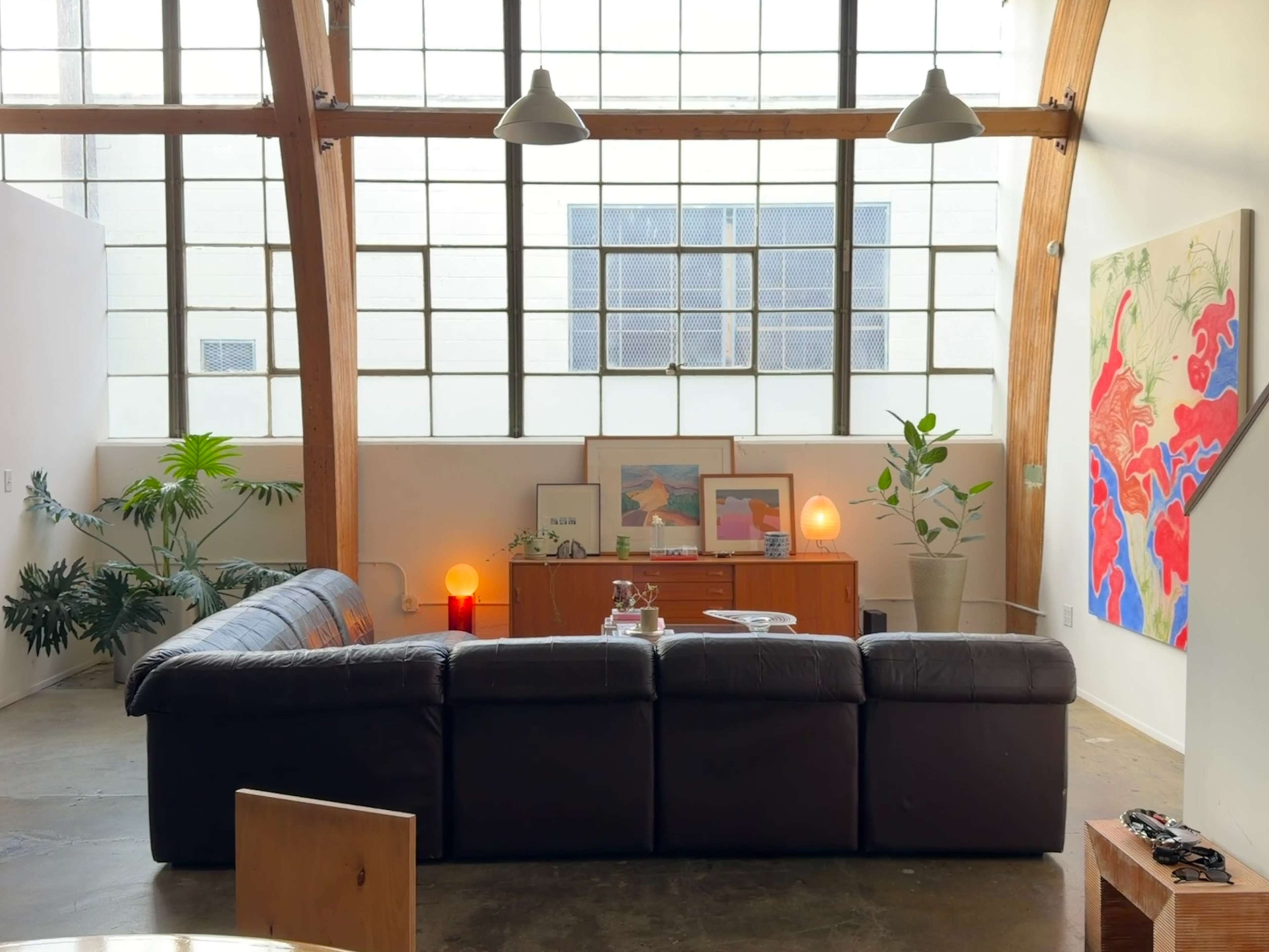 The image shows a spacious living area with large windows, a dark sofa facing a wooden cabinet, and artwork displayed on the walls.