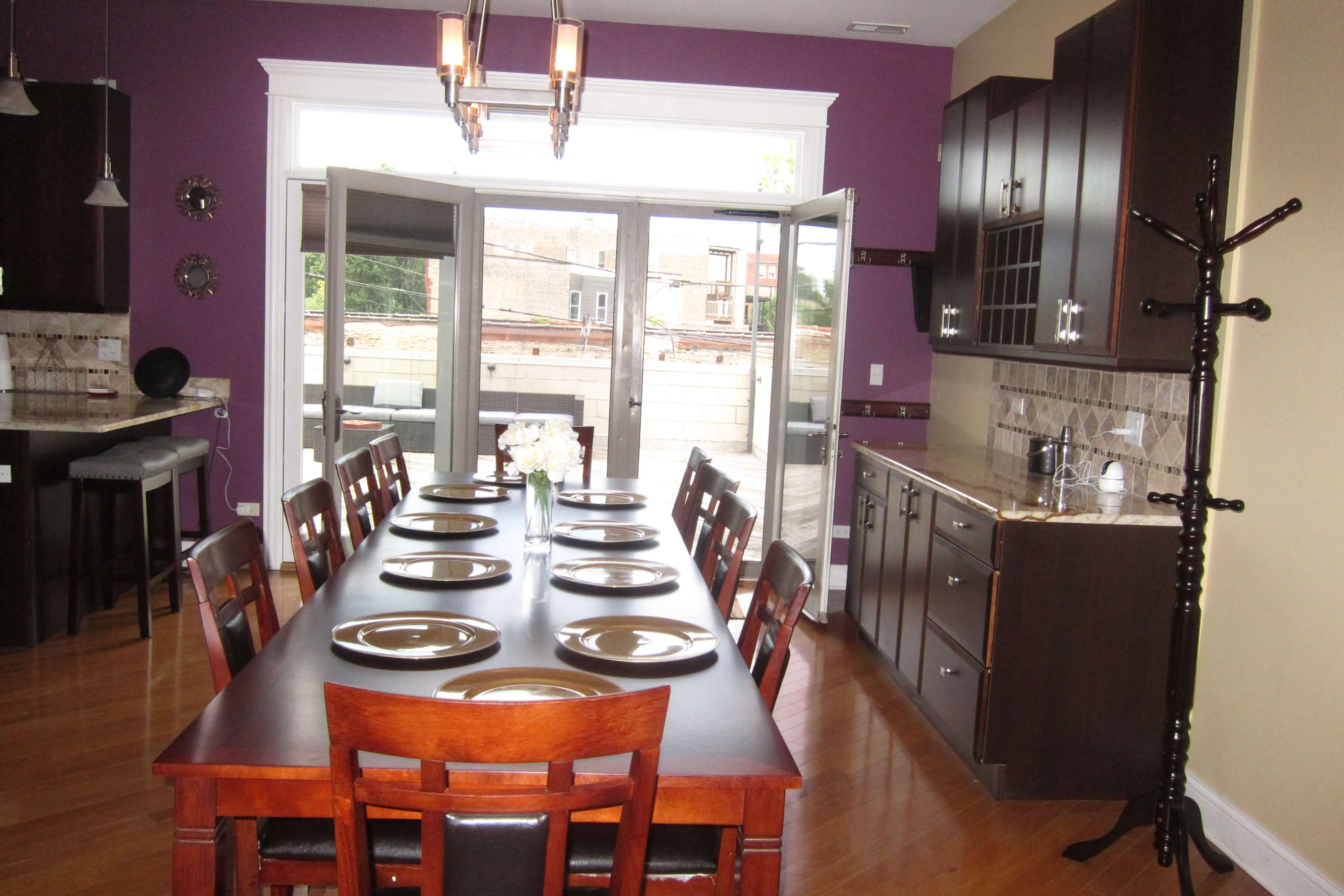A long wooden dining table with plates set is positioned in a brightly lit kitchen, featuring purple walls and large double doors that open to an outdoor area.