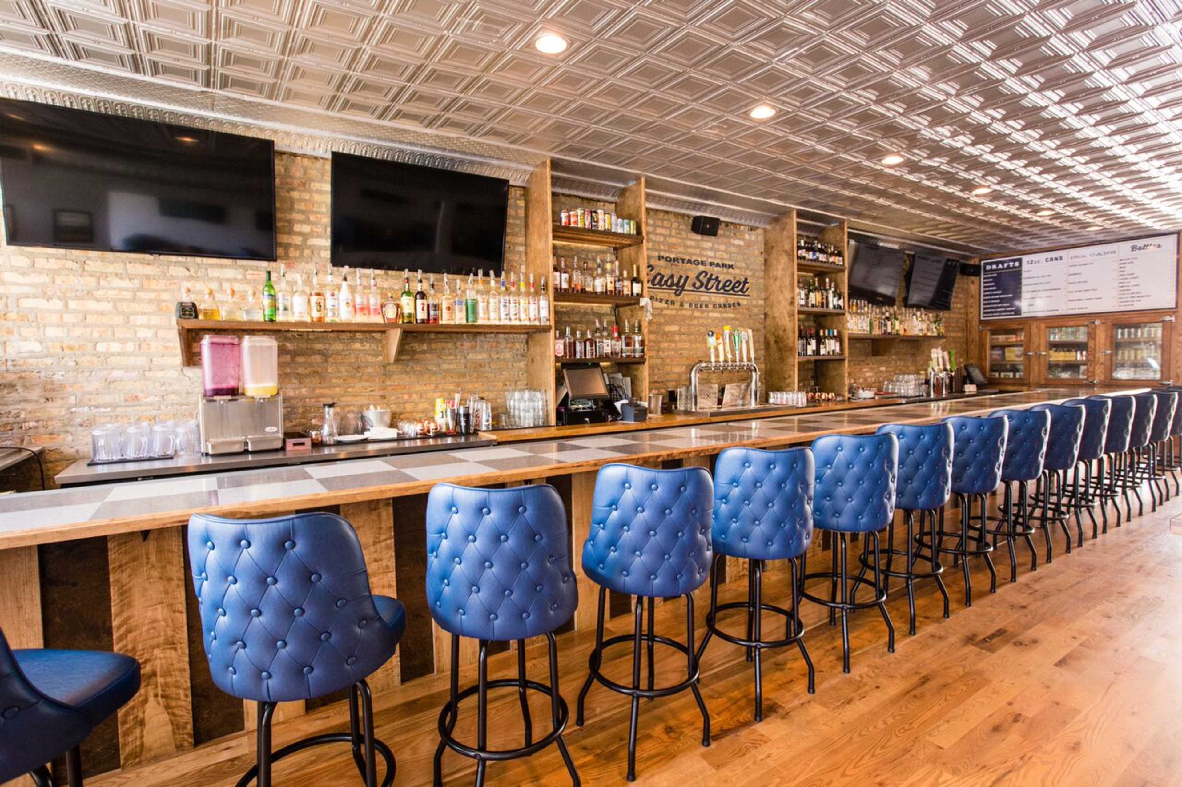 The image shows a modern bar with a long wooden counter, blue tufted bar stools, and various bottles displayed on shelves against a brick wall.