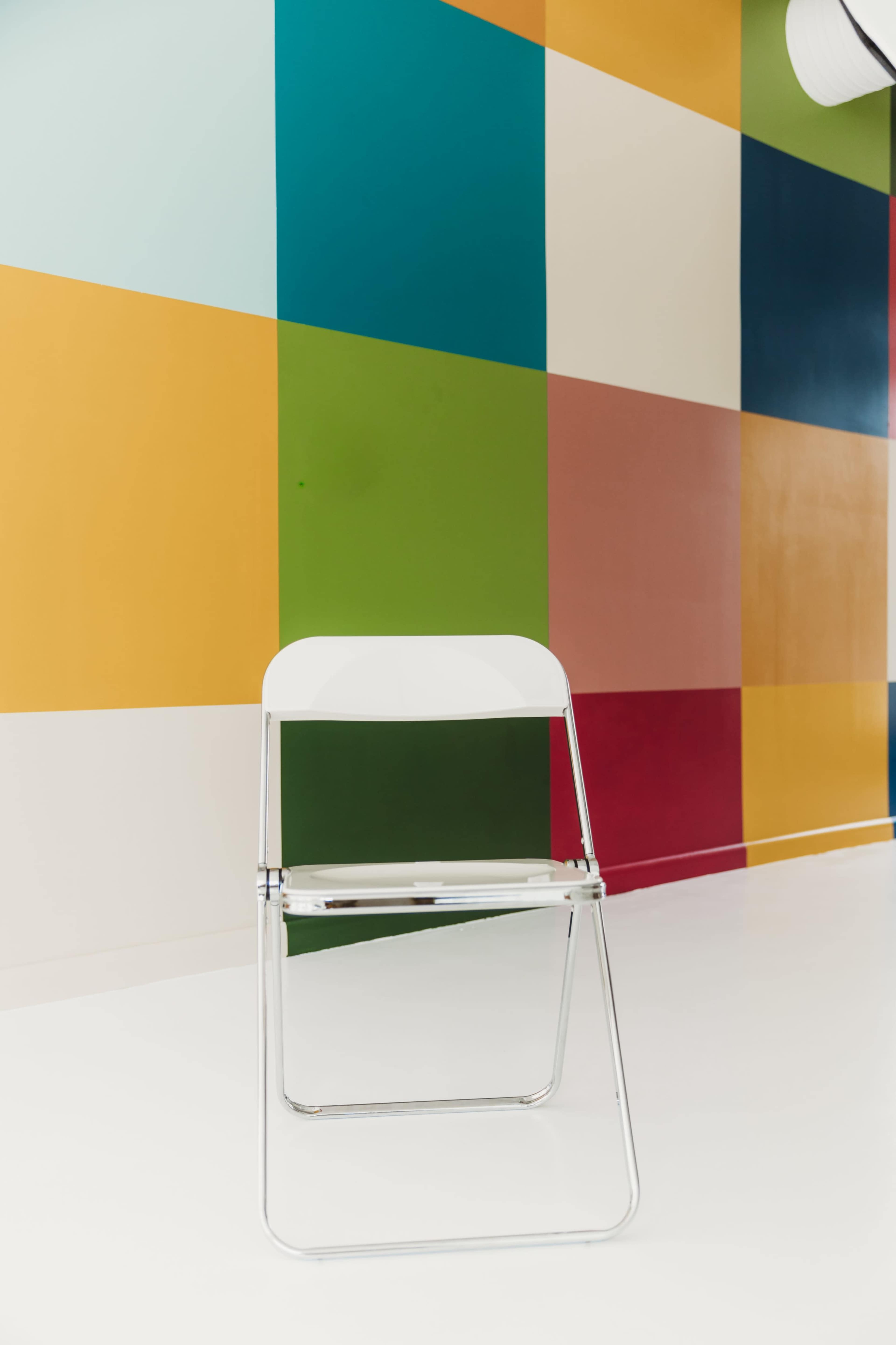A white folding chair is positioned against a brightly colored patchwork wall.