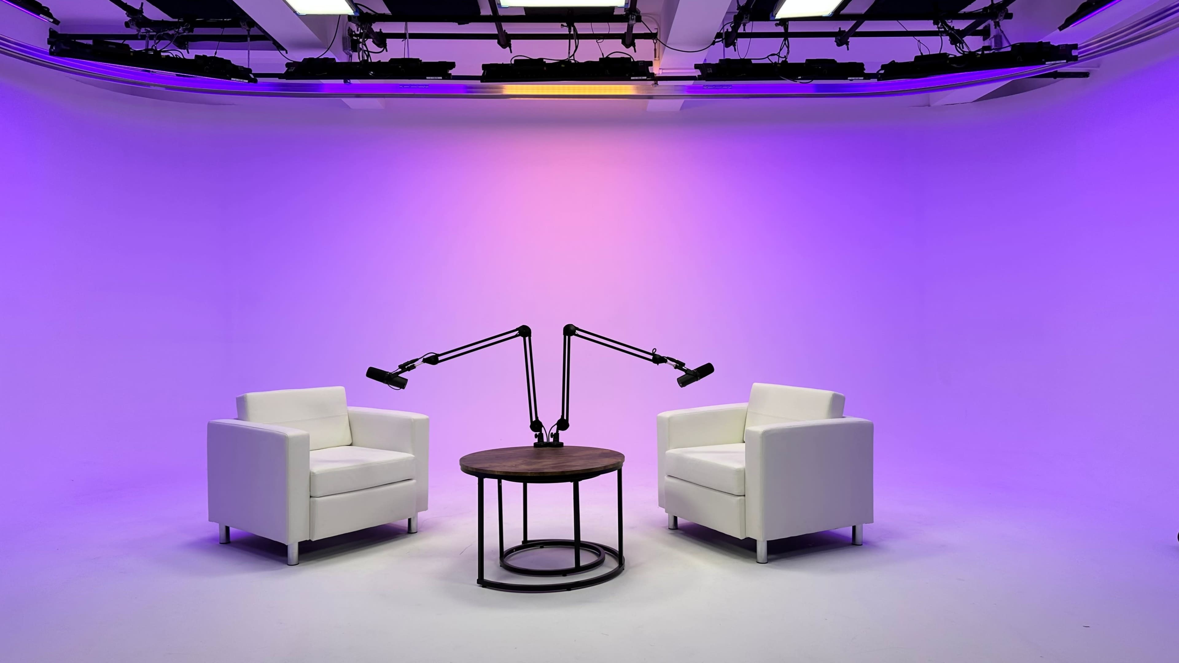A minimalist studio setup features two white chairs positioned around a round wooden table, illuminated by adjustable lamps against a purple backdrop.
