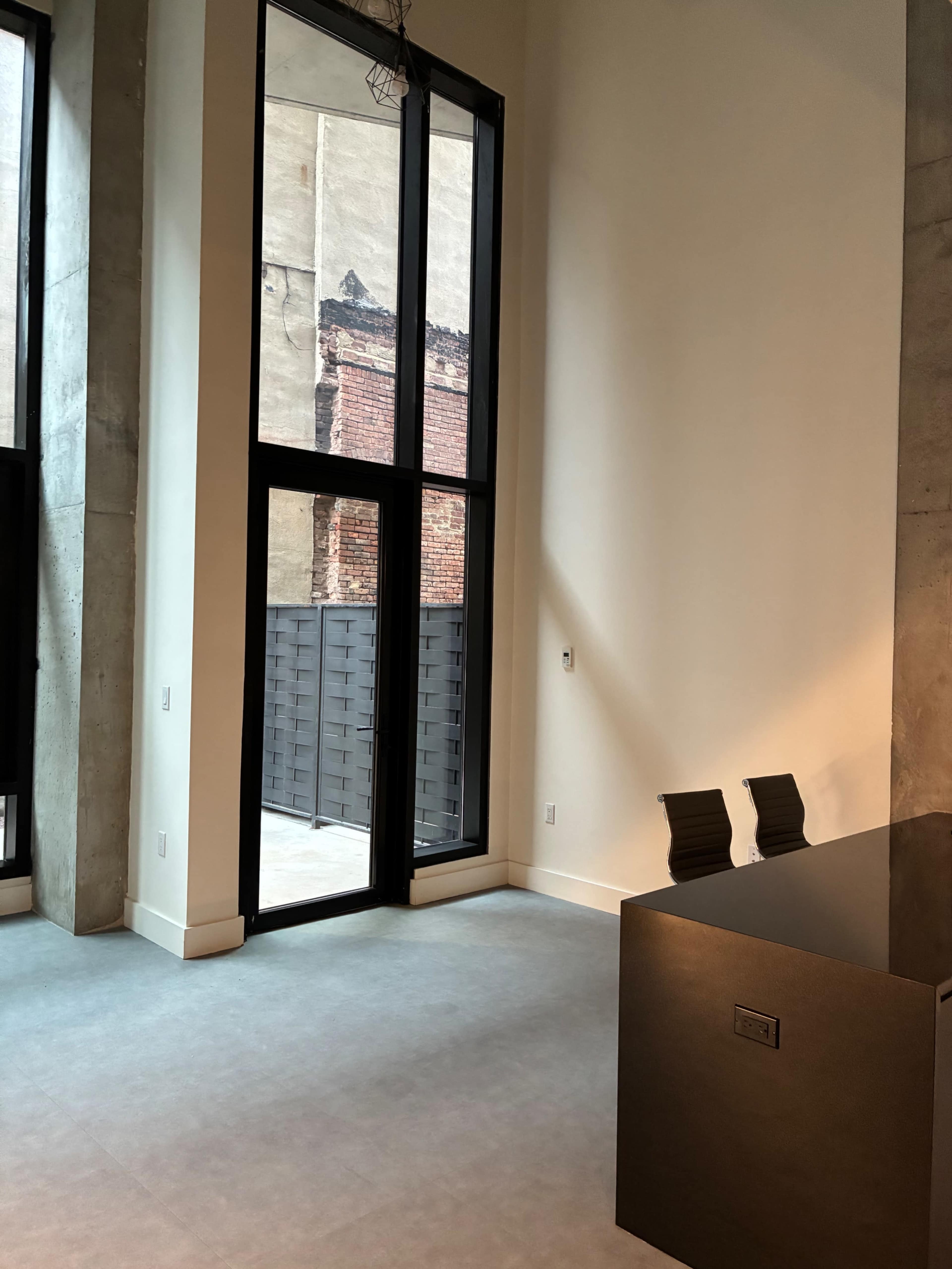 The image shows a spacious interior with large windows revealing a brick wall outside, featuring a minimalist desk and two chairs.