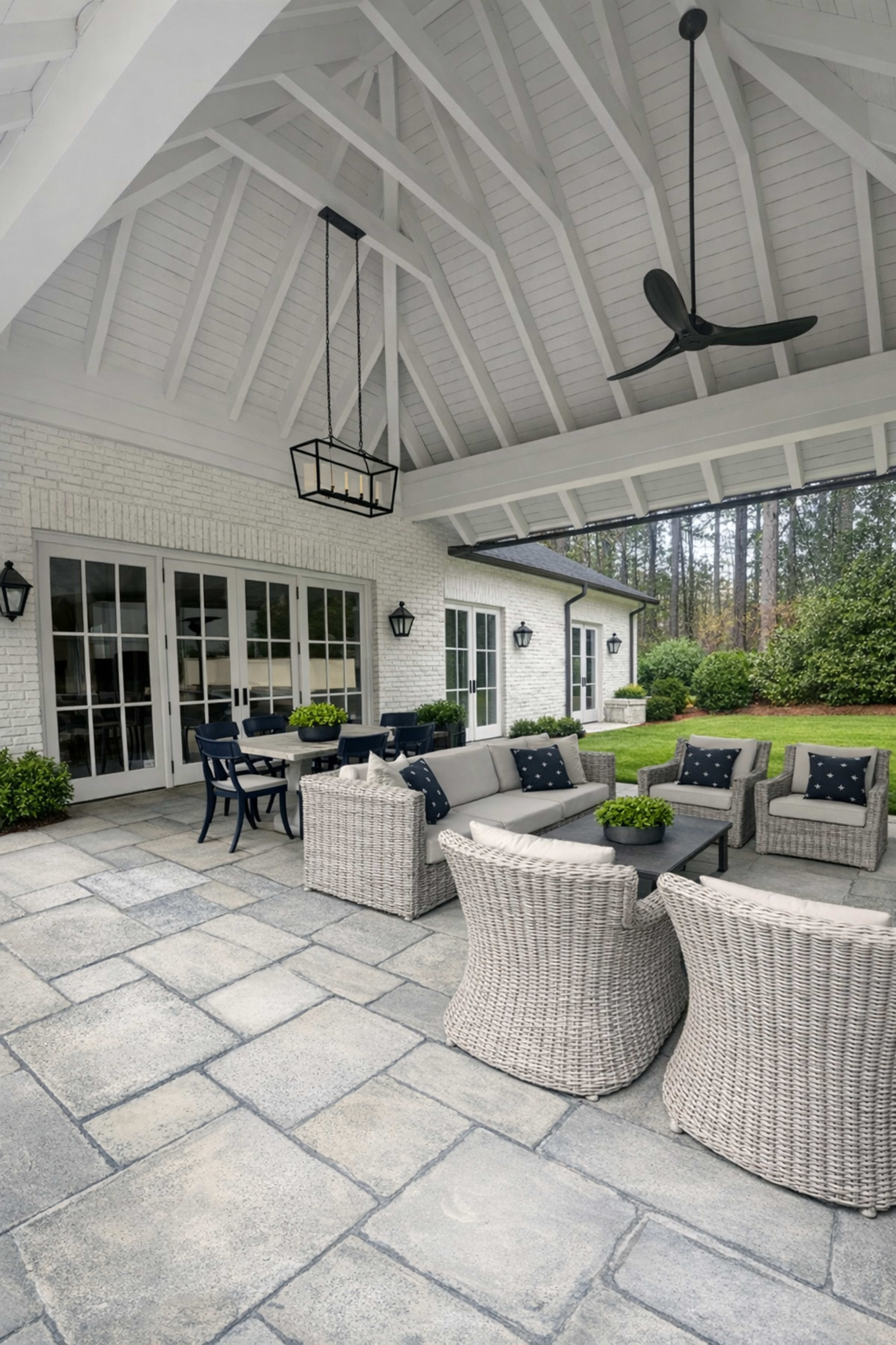 The image shows a covered patio with a seating area featuring gray wicker furniture and a dining table, surrounded by greenery and stone flooring.