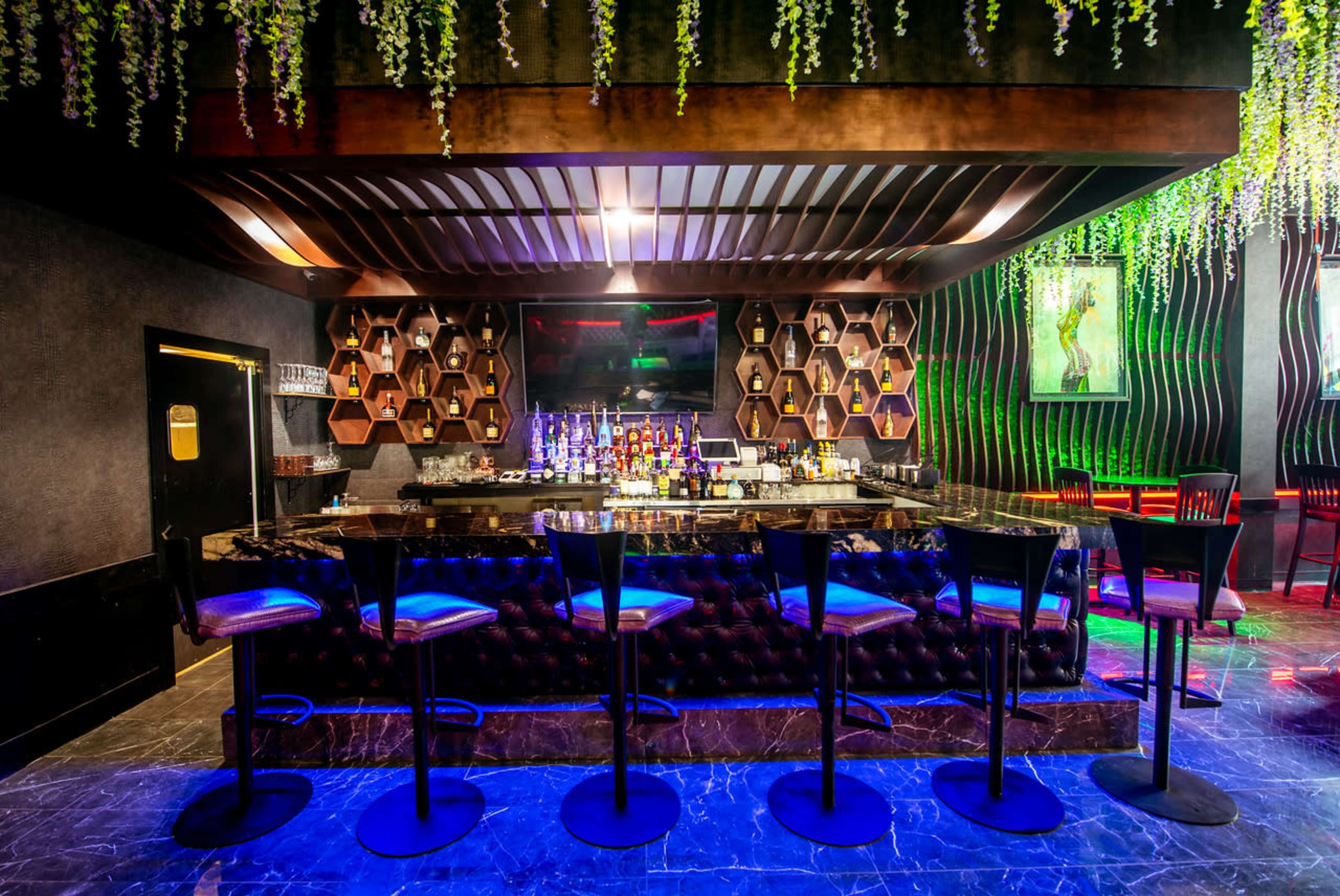 A modern bar with a hexagonal-patterned back wall, dark wooden accents, and neon lighting illuminating the seating area.