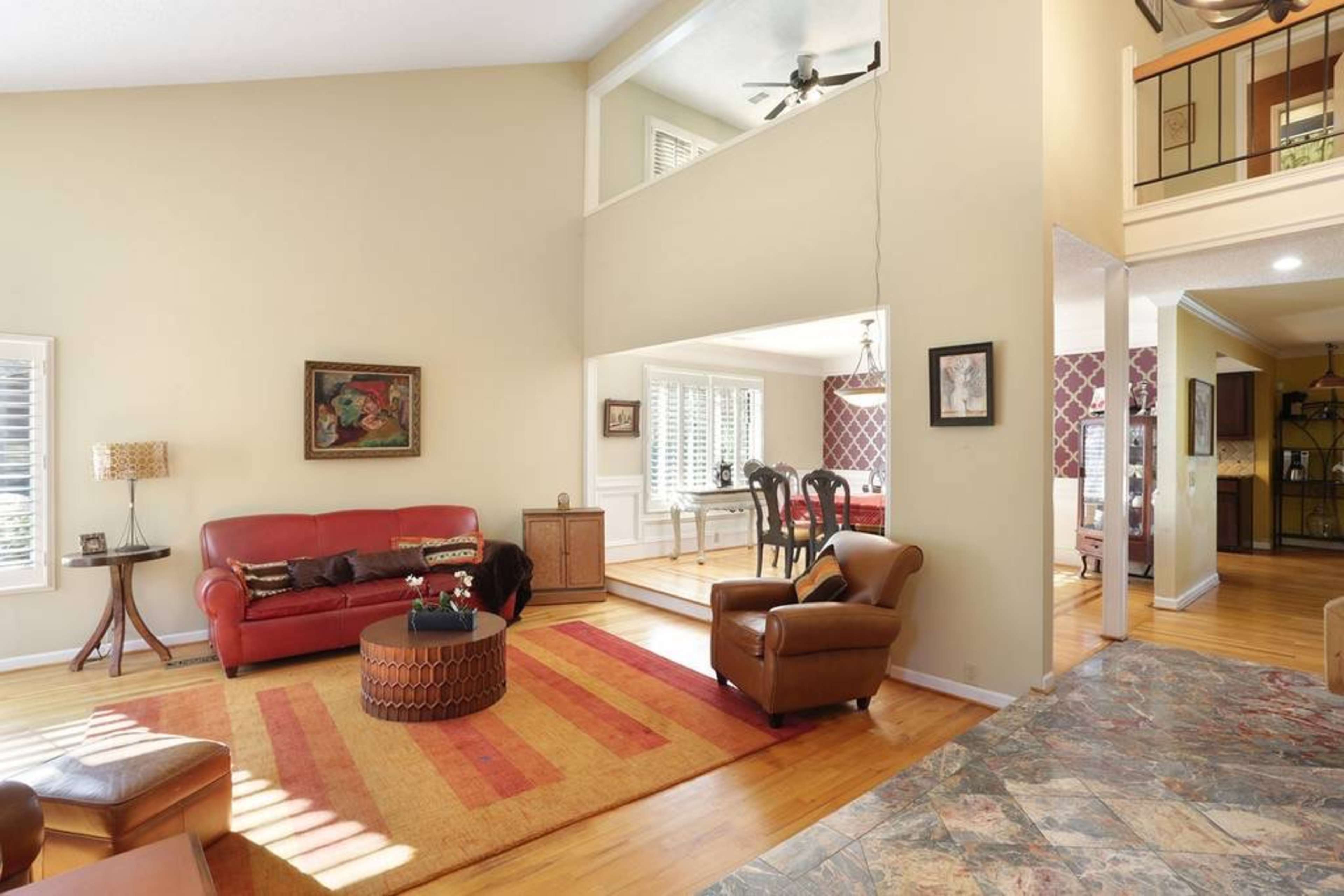 The image shows a spacious living room furnished with a red sofa, a brown armchair, a wooden coffee table, and a large area rug, featuring a view into a dining area with a table set for multiple people.