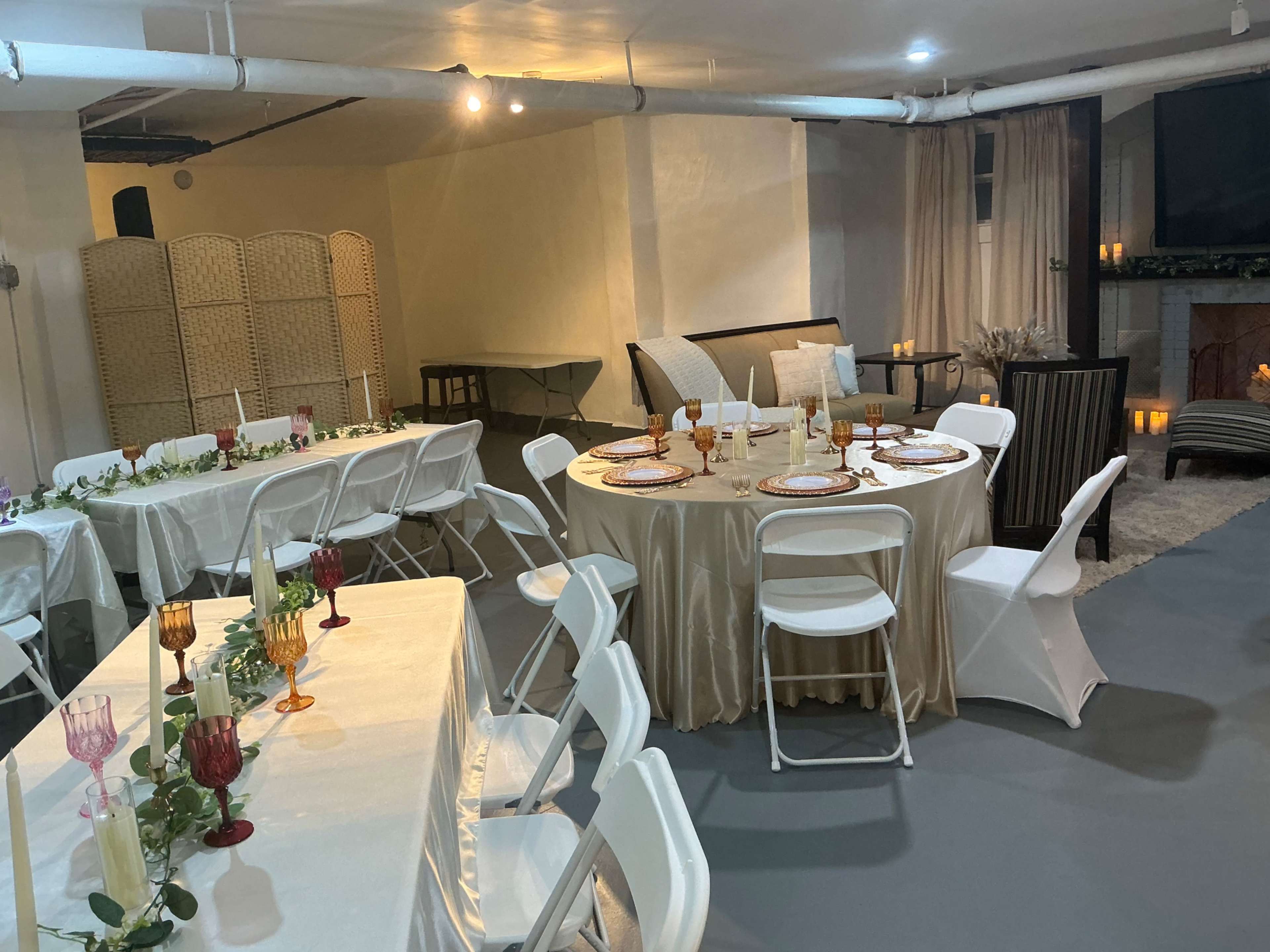 The image shows a dimly lit indoor event space set up with multiple tables, each elegantly decorated with tableware and centerpieces, creating an inviting atmosphere for a gathering.