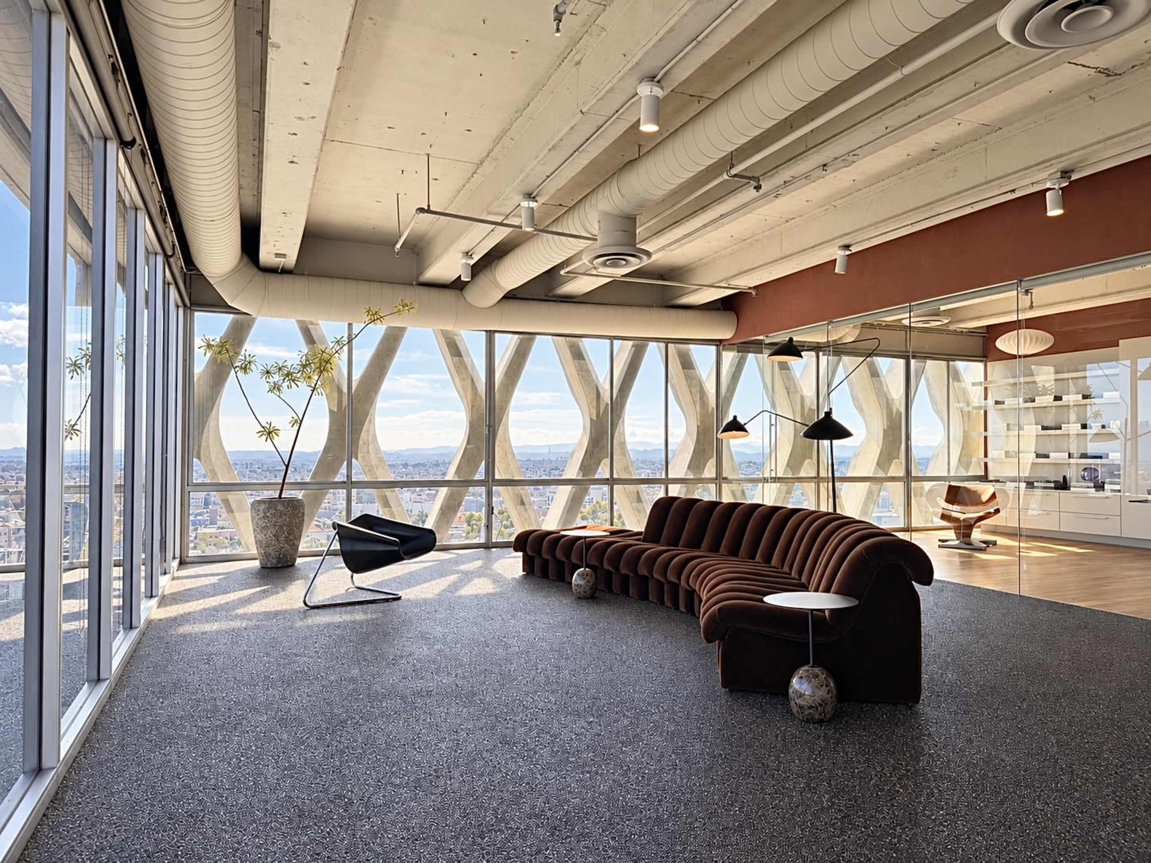 The image shows a modern interior space with a curved brown sofa, a black chair, and large windows offering a view of the city skyline.