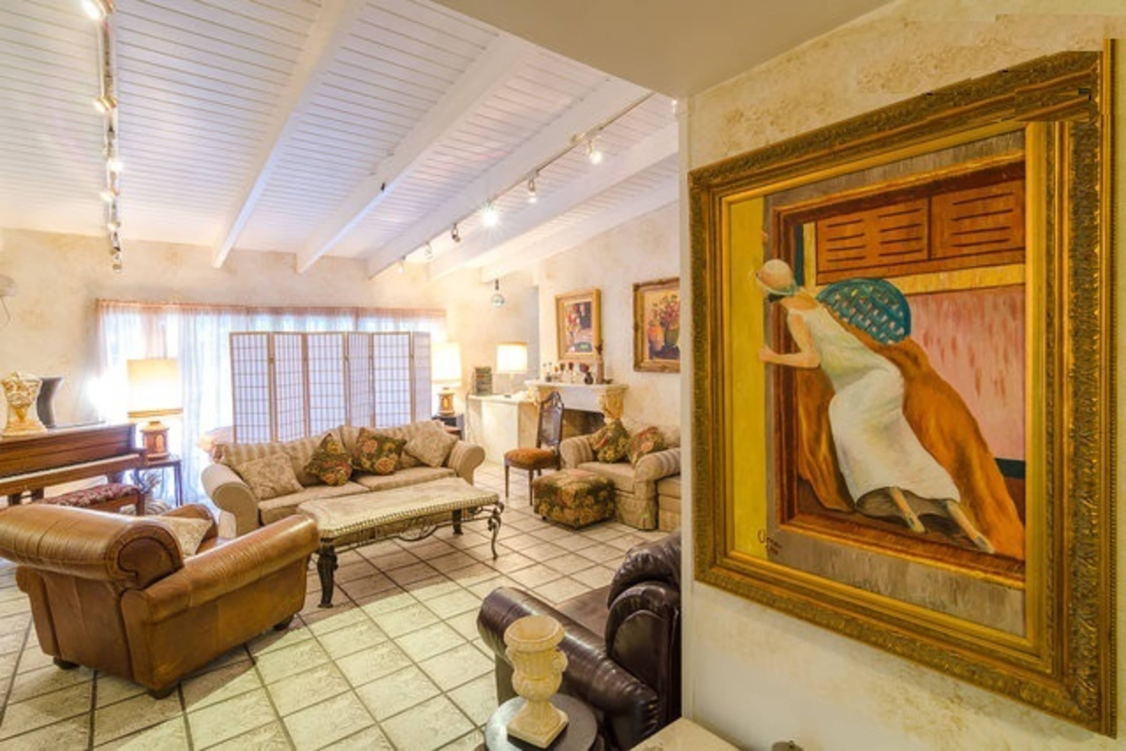 A spacious living room features a mix of upholstered furniture, a decorative painting, and large windows allowing natural light to filter in.