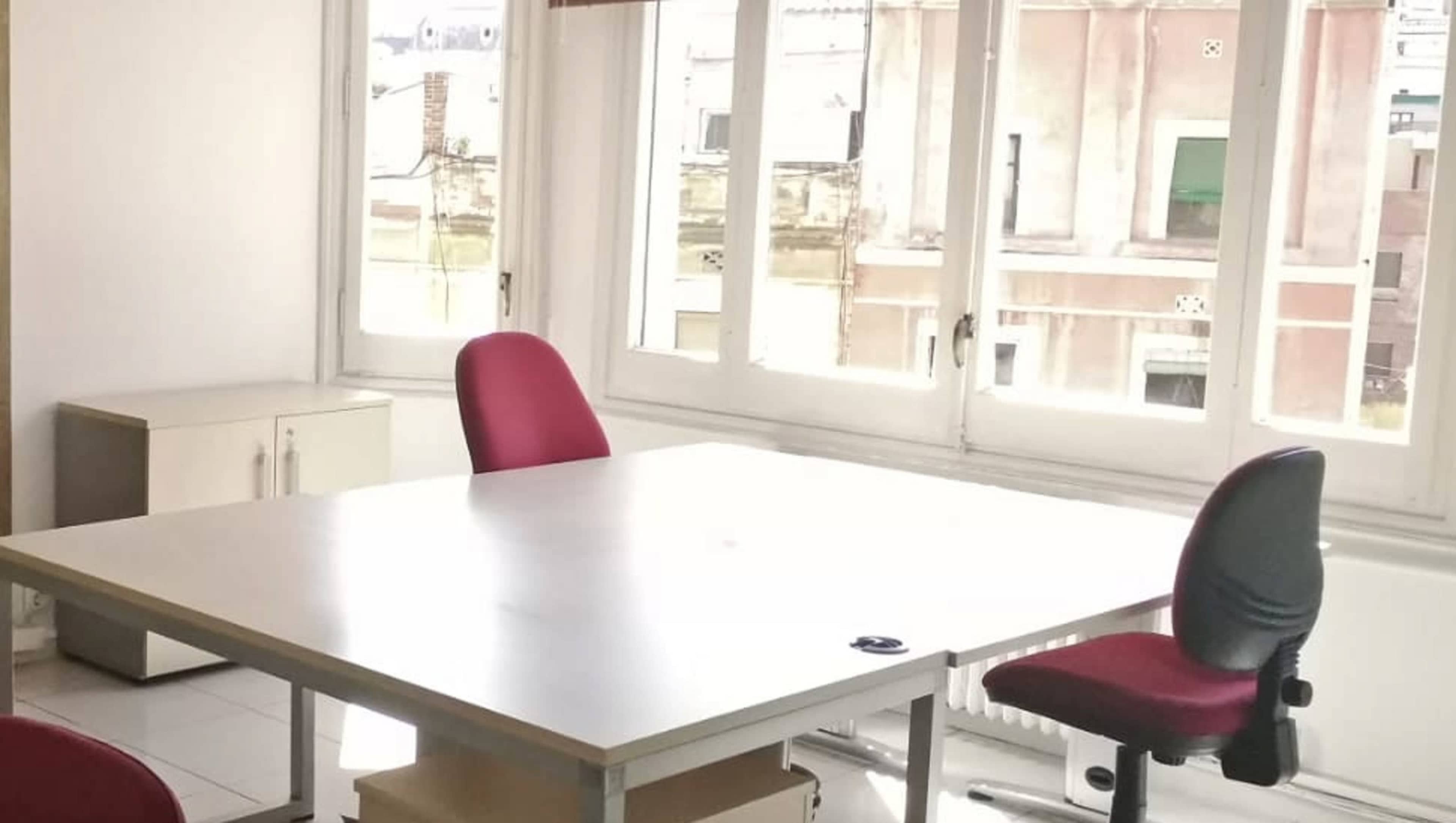 A bright office space features a large white table and two red chairs, with windows offering a view of neighboring buildings.