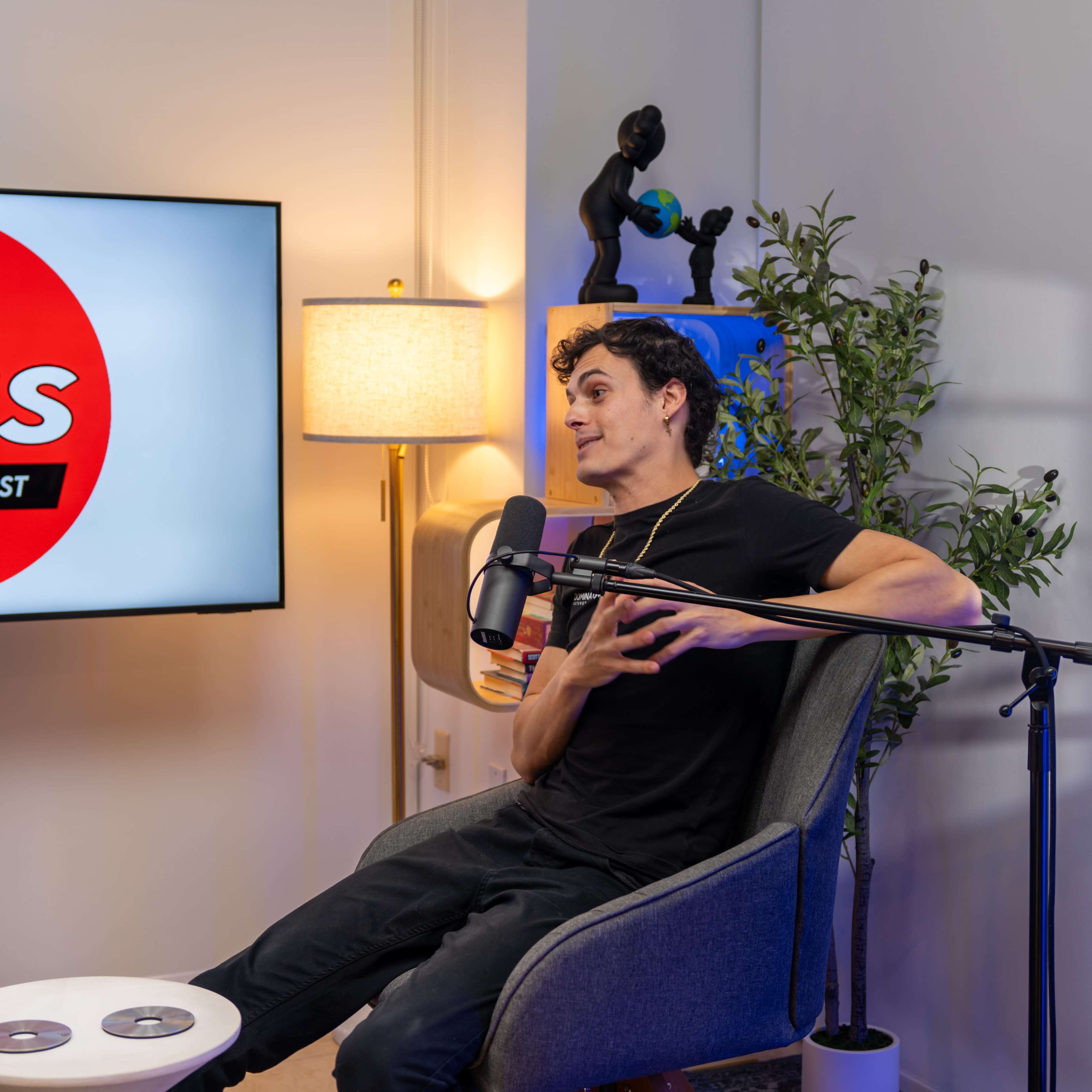 A young man sits in a chair with a microphone in front of him, gesturing as he speaks during a podcast recording in a well-lit studio.