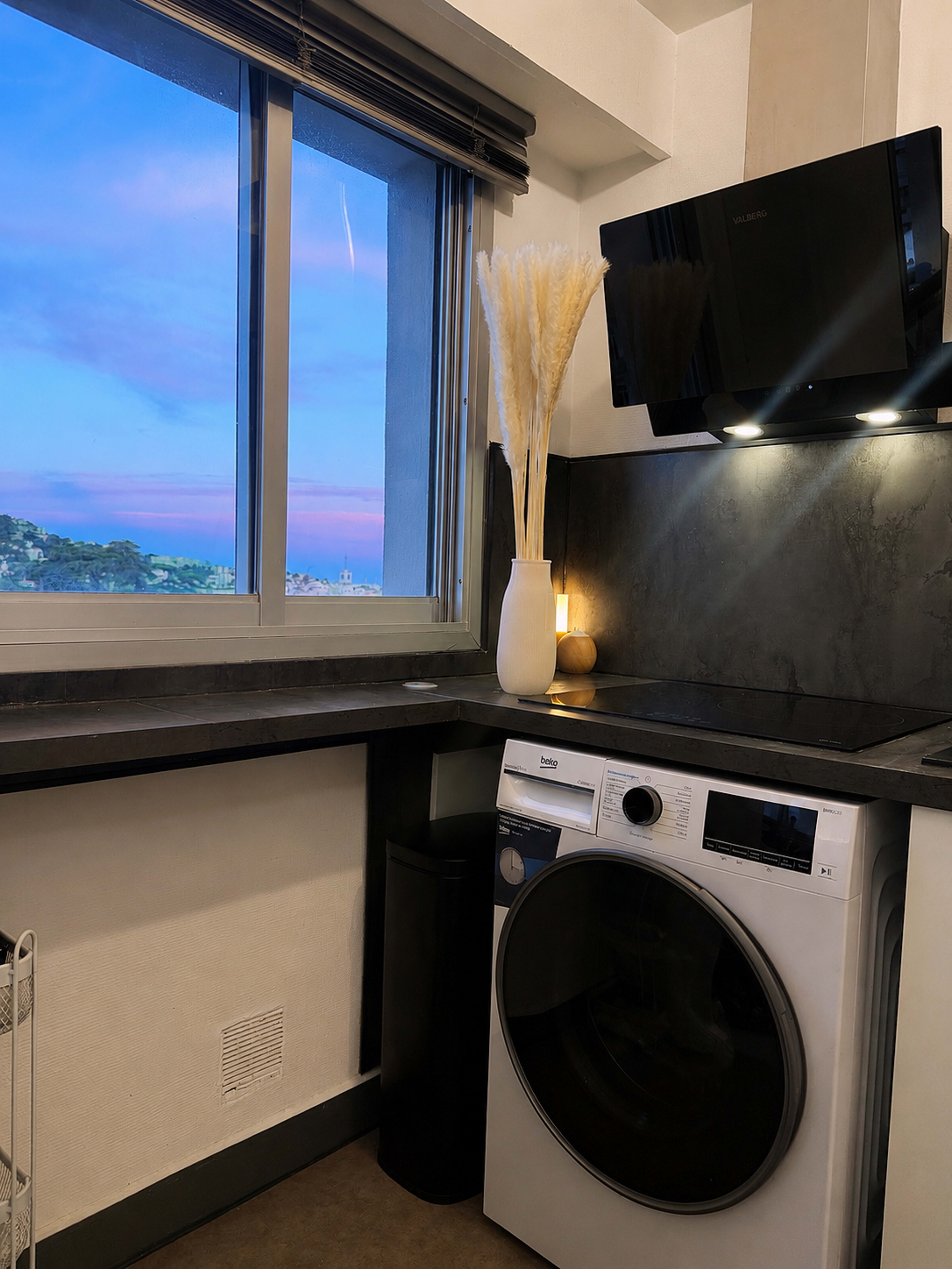 A modern laundry room features a washing machine beside a counter with a window overlooking a scenic view, decorated with a vase of dried pampas grass and a candle.