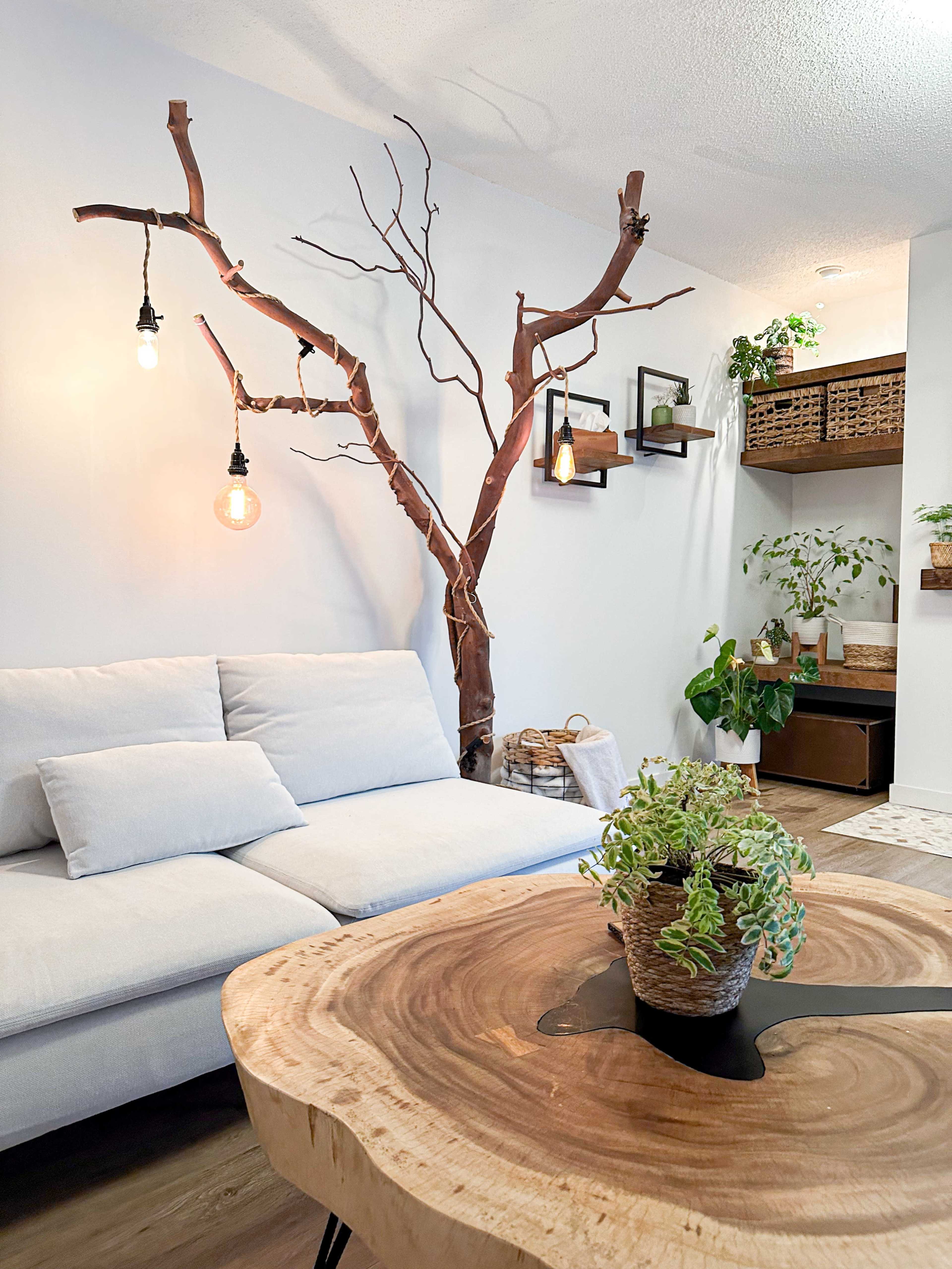 A modern living room features a light gray sofa, a round wooden coffee table, and a sculptural tree branch mounted on the wall with hanging light fixtures.