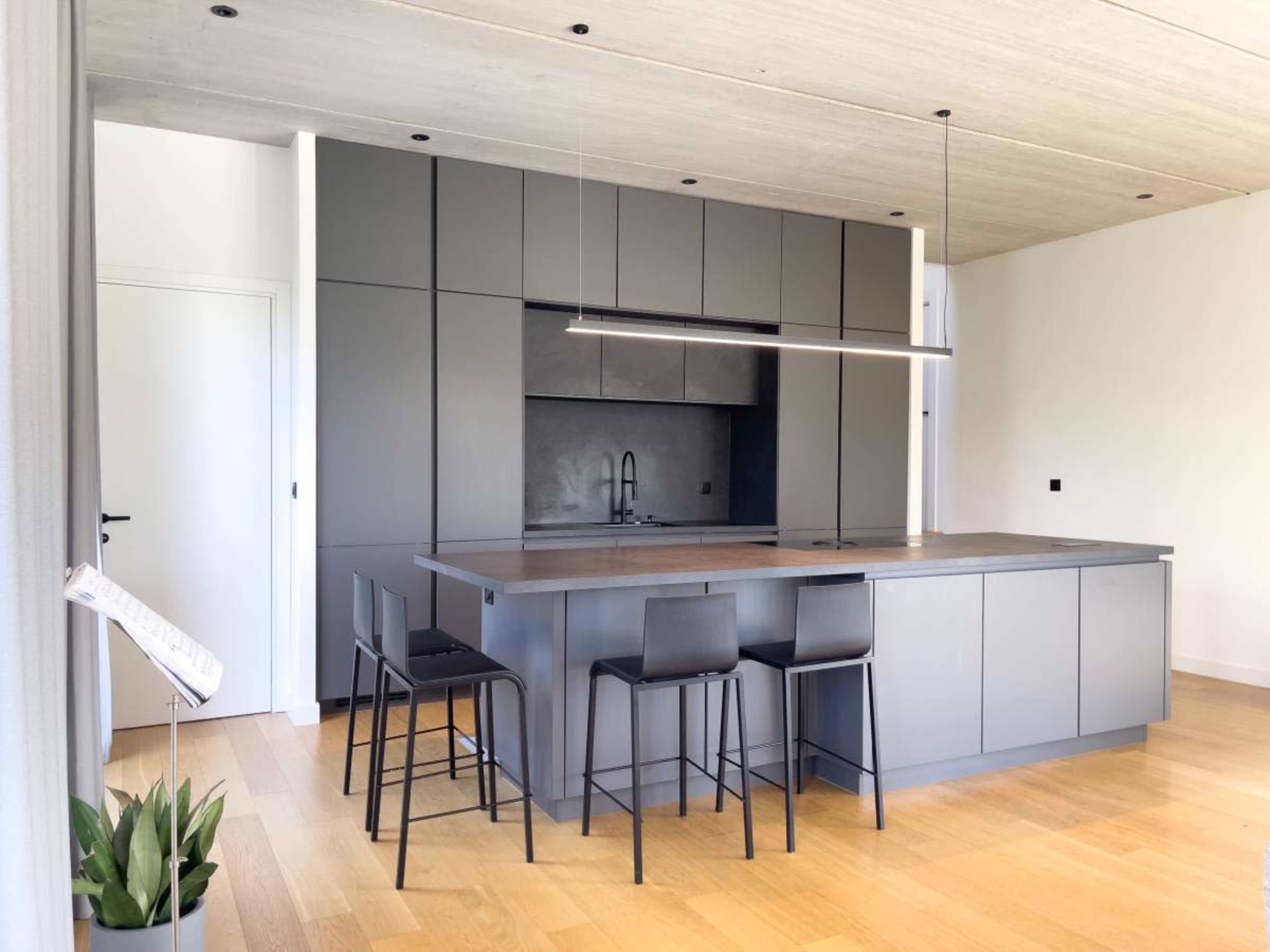 The image depicts a modern kitchen featuring a gray island with high stools, a minimalist design, and wood flooring.