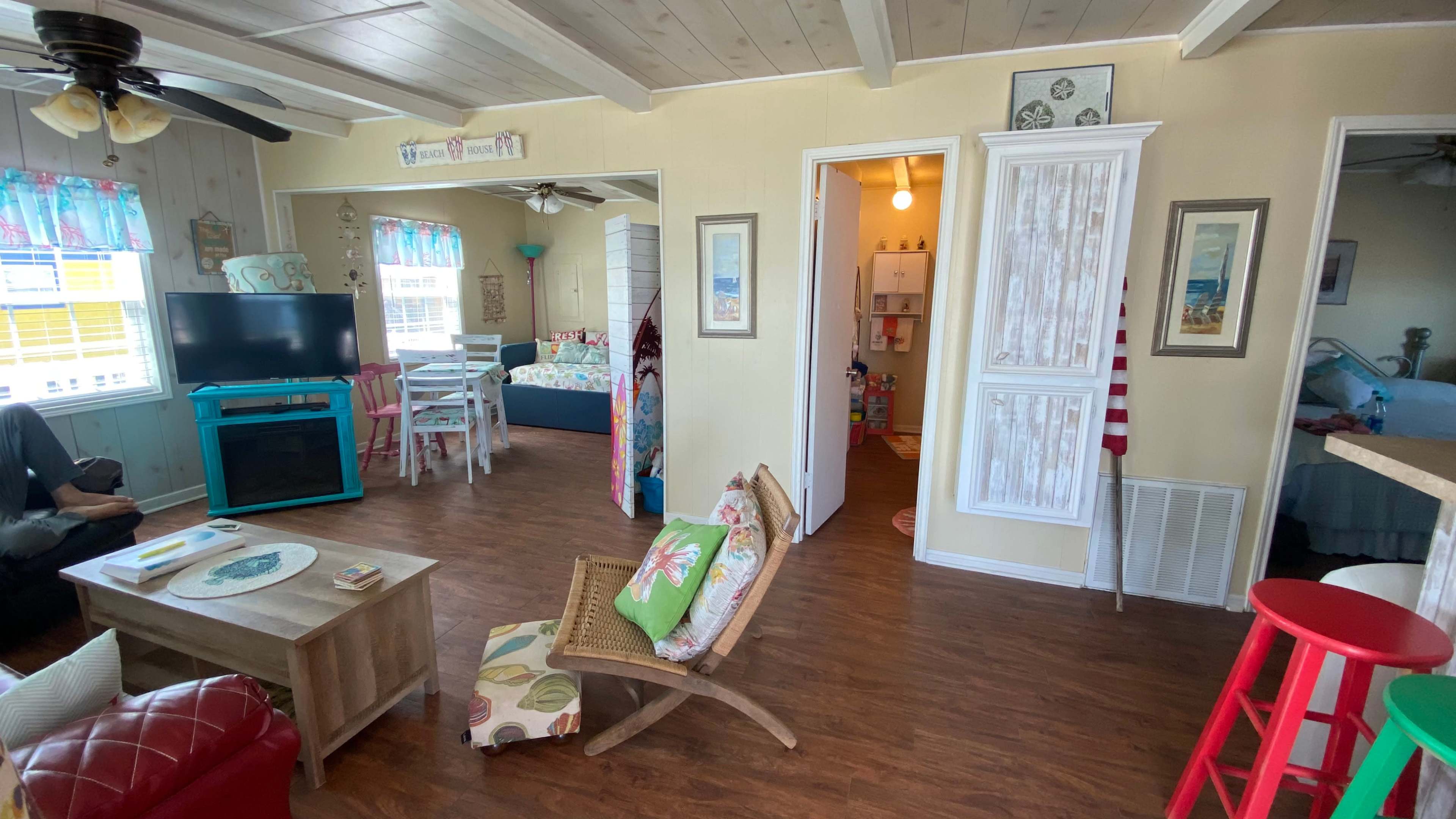 The image shows a bright, open living space featuring a seating area with colorful furnishings, a TV, and a view into a bedroom through an adjacent doorway.