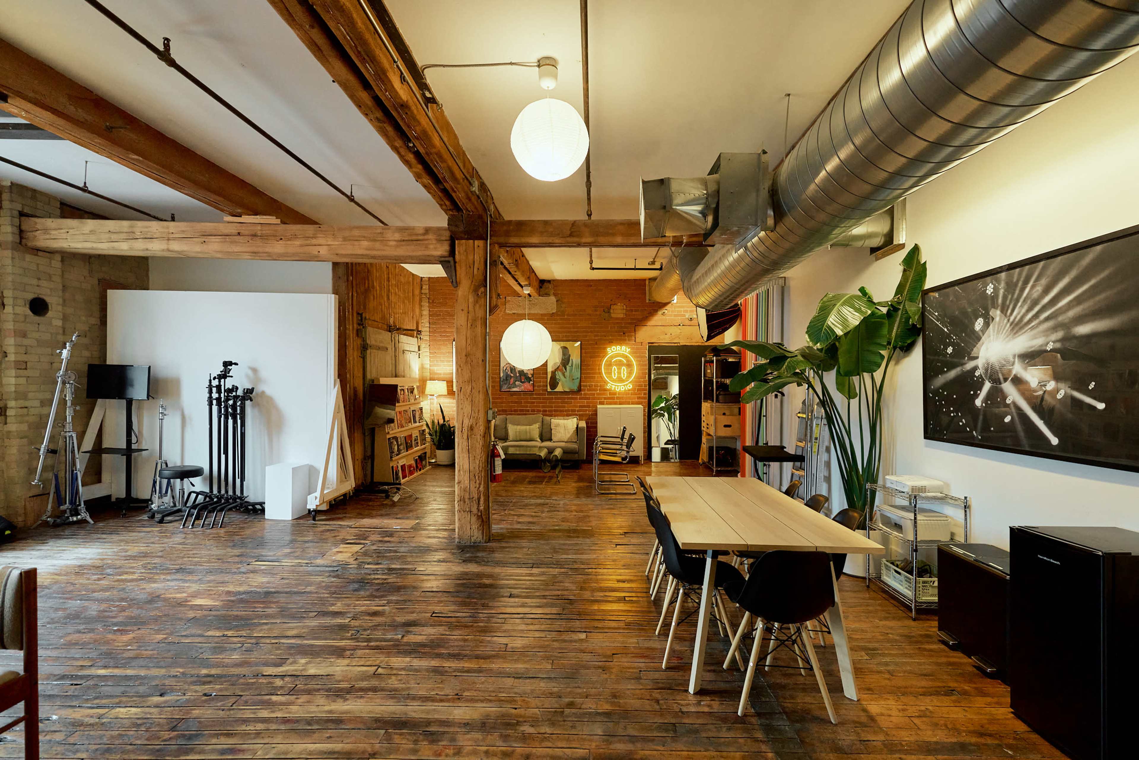 The image shows a spacious, modern studio with wooden floors, exposed beams, and various seating areas, including a long table and lounge area.