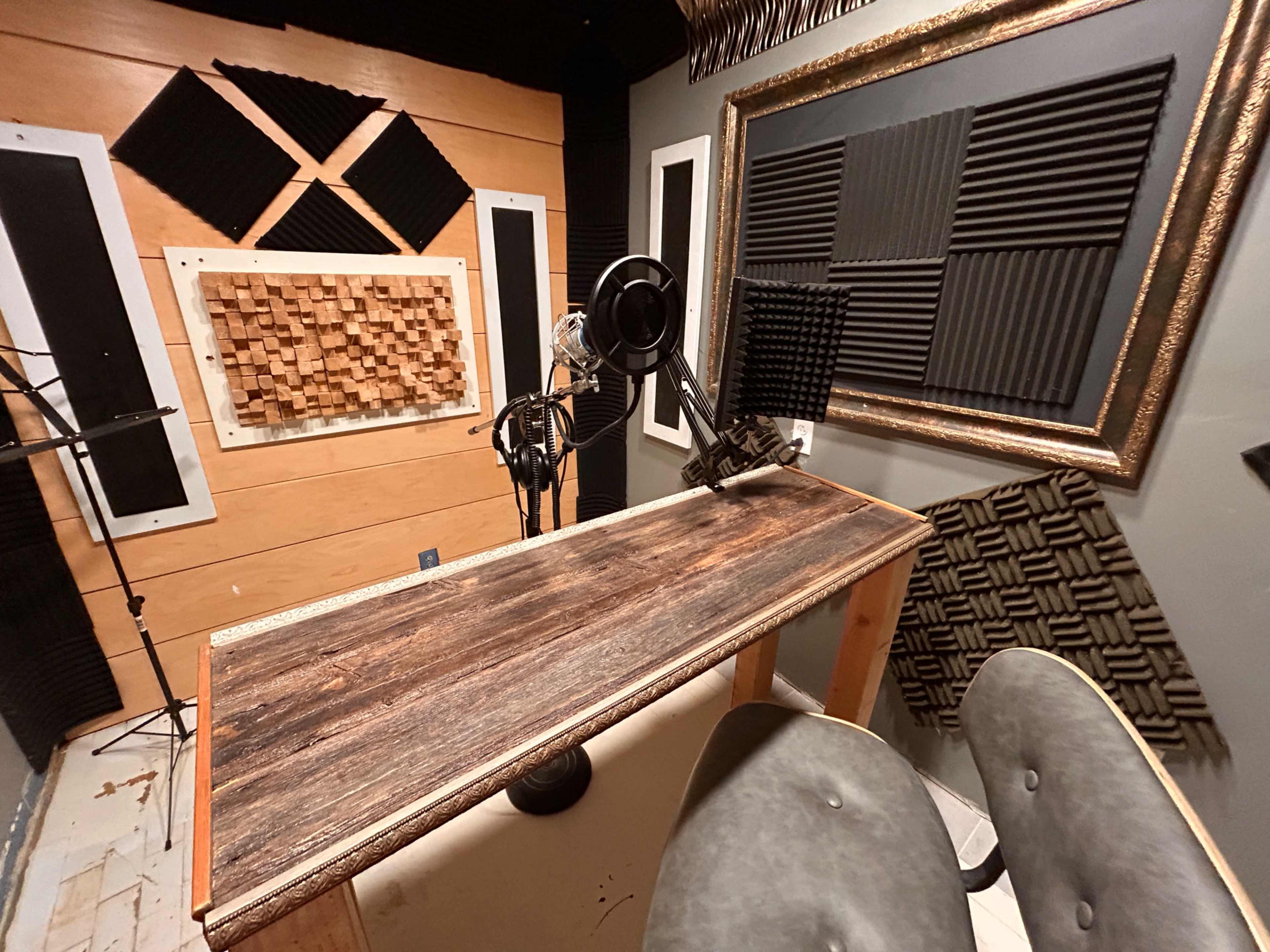 The image shows a small recording studio with a wooden desk, two chairs, and soundproofing panels on the walls.