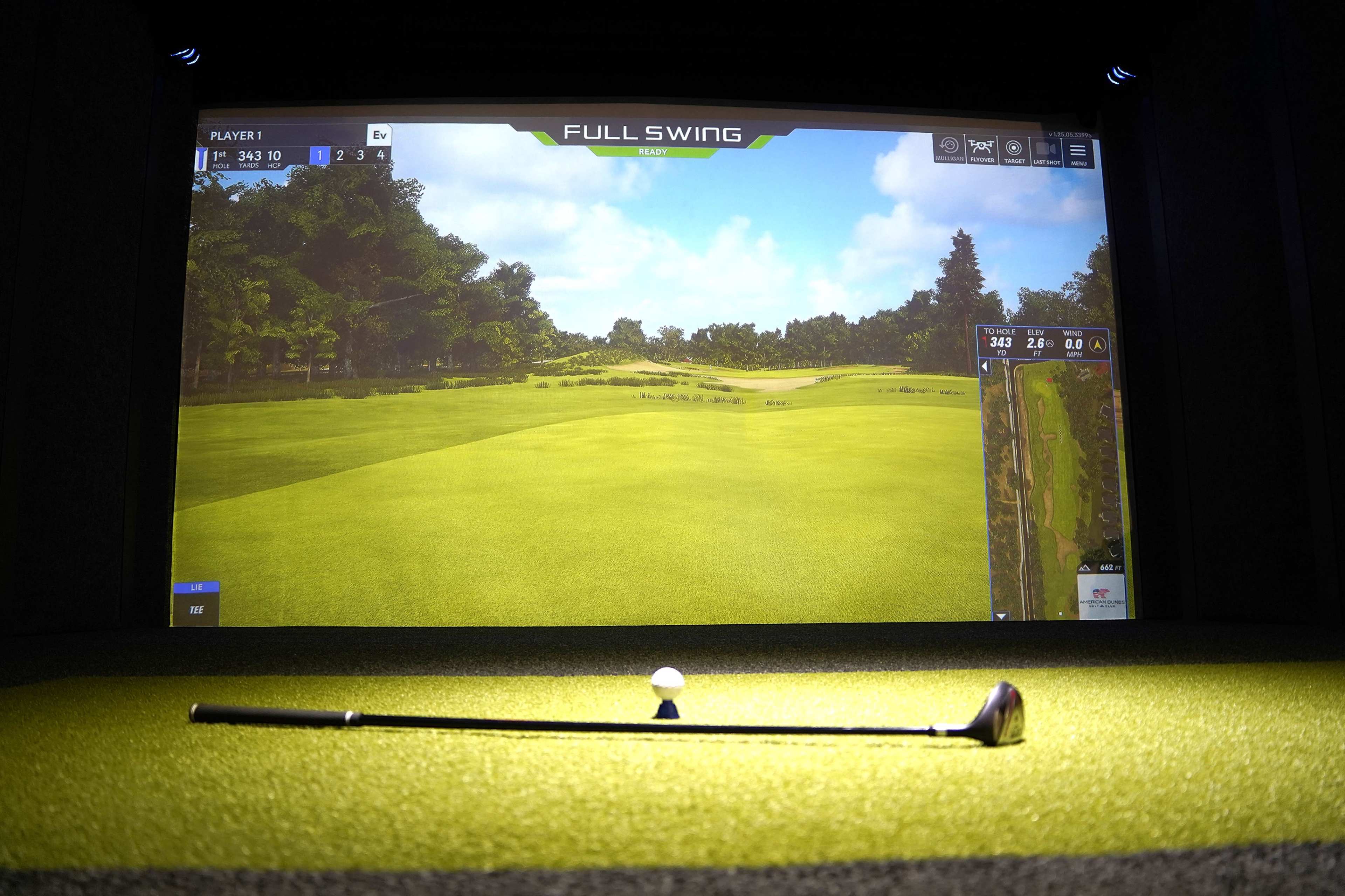 The image shows a golf simulator setup with a club and golf ball in the foreground and a large screen displaying a virtual golf course in the background.