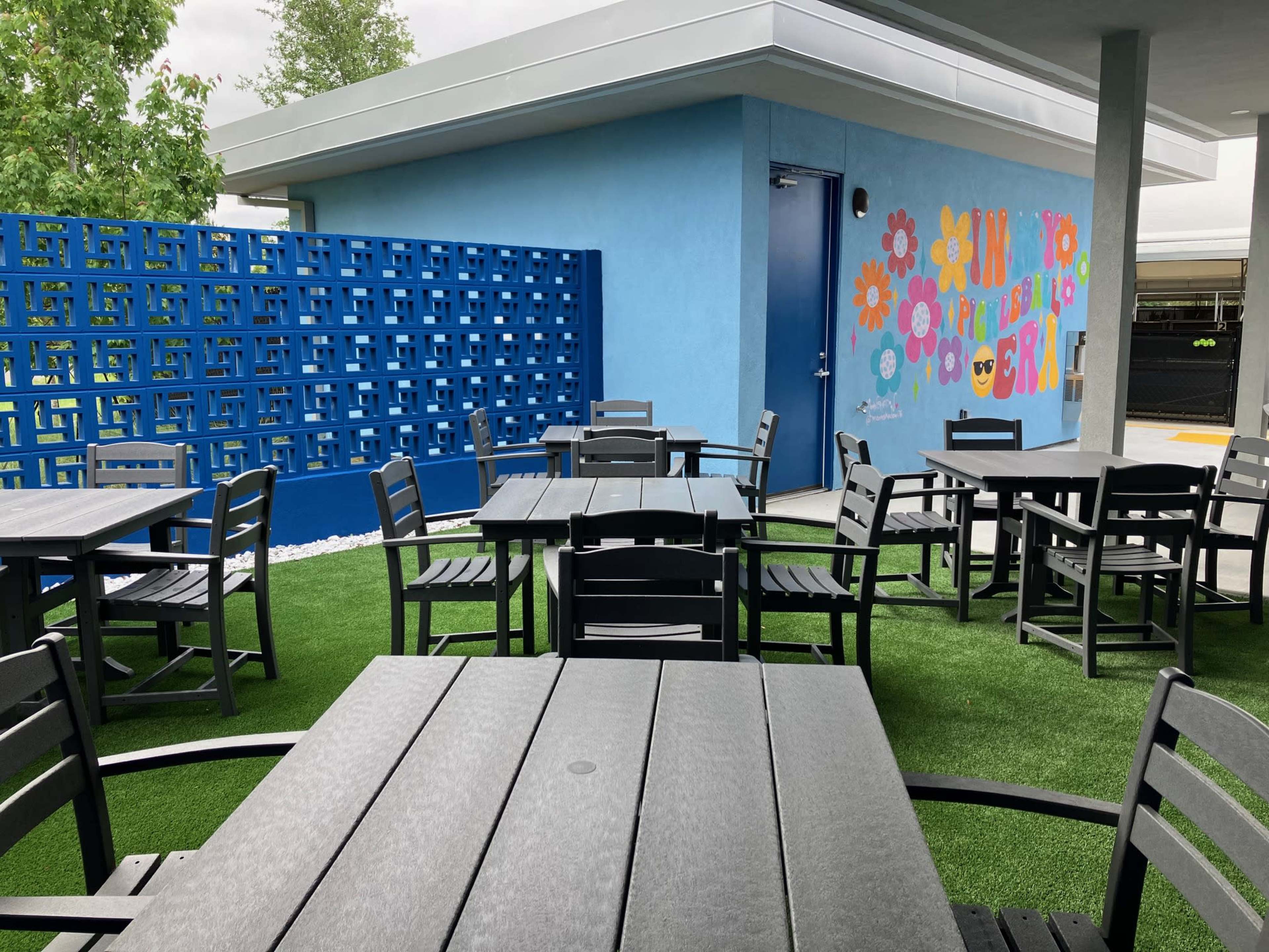 The image shows an outdoor dining area with black tables and chairs set on green turf, alongside a blue wall featuring colorful flower decals.
