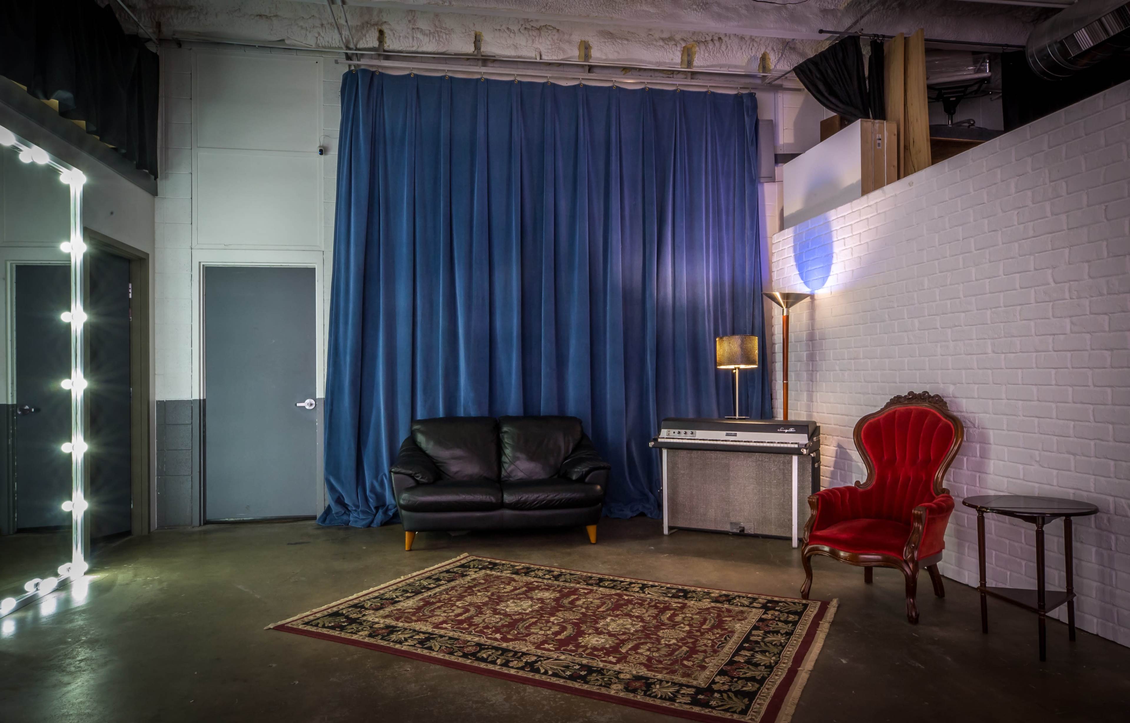 A studio room with a dark blue curtain, a black leather couch, a red armchair, a side table, and a vintage keyboard on a gray surface.