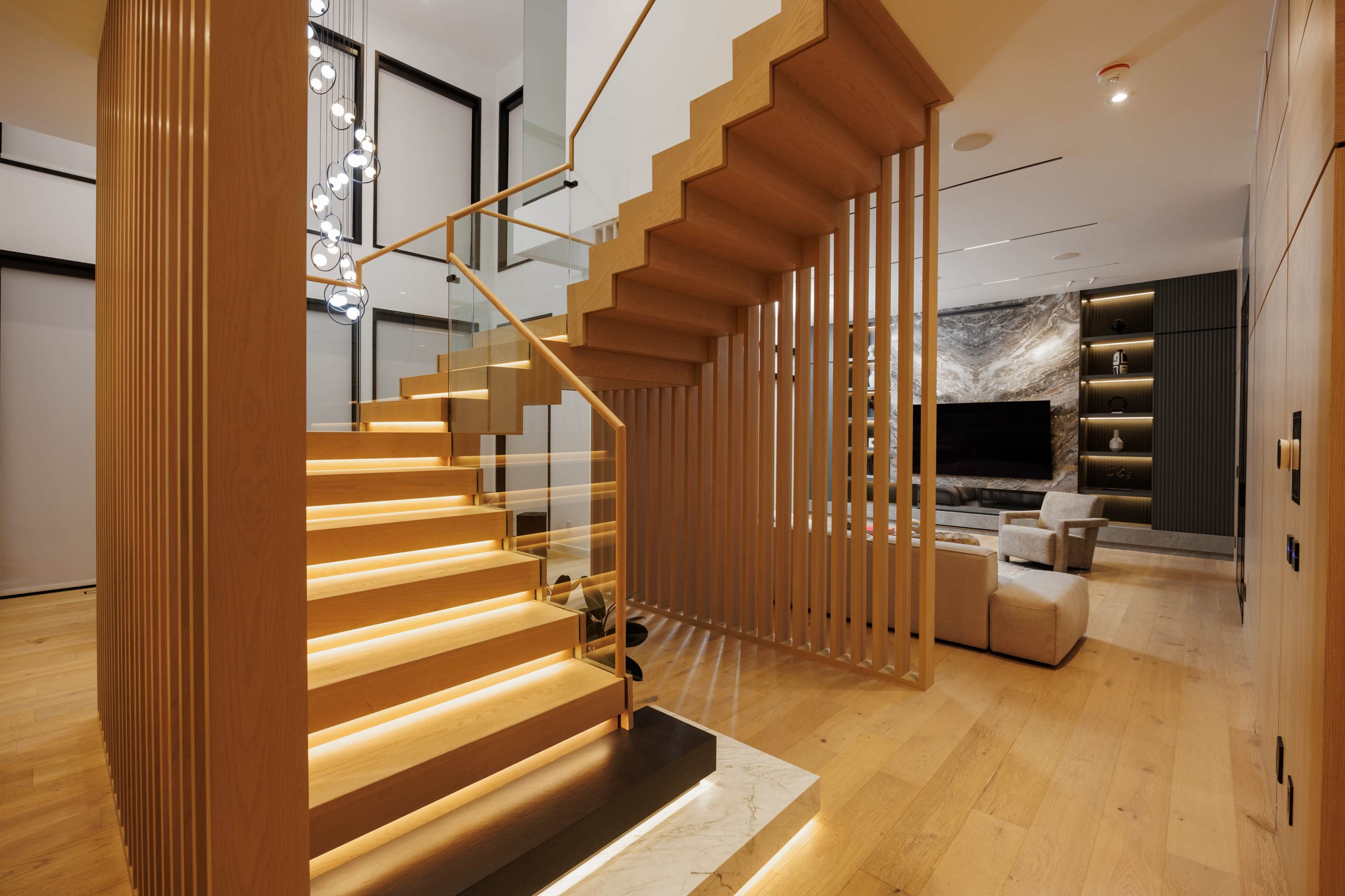 The image shows a modern interior with a wooden staircase illuminated by lights, leading to an open living area with a fireplace and contemporary furniture.