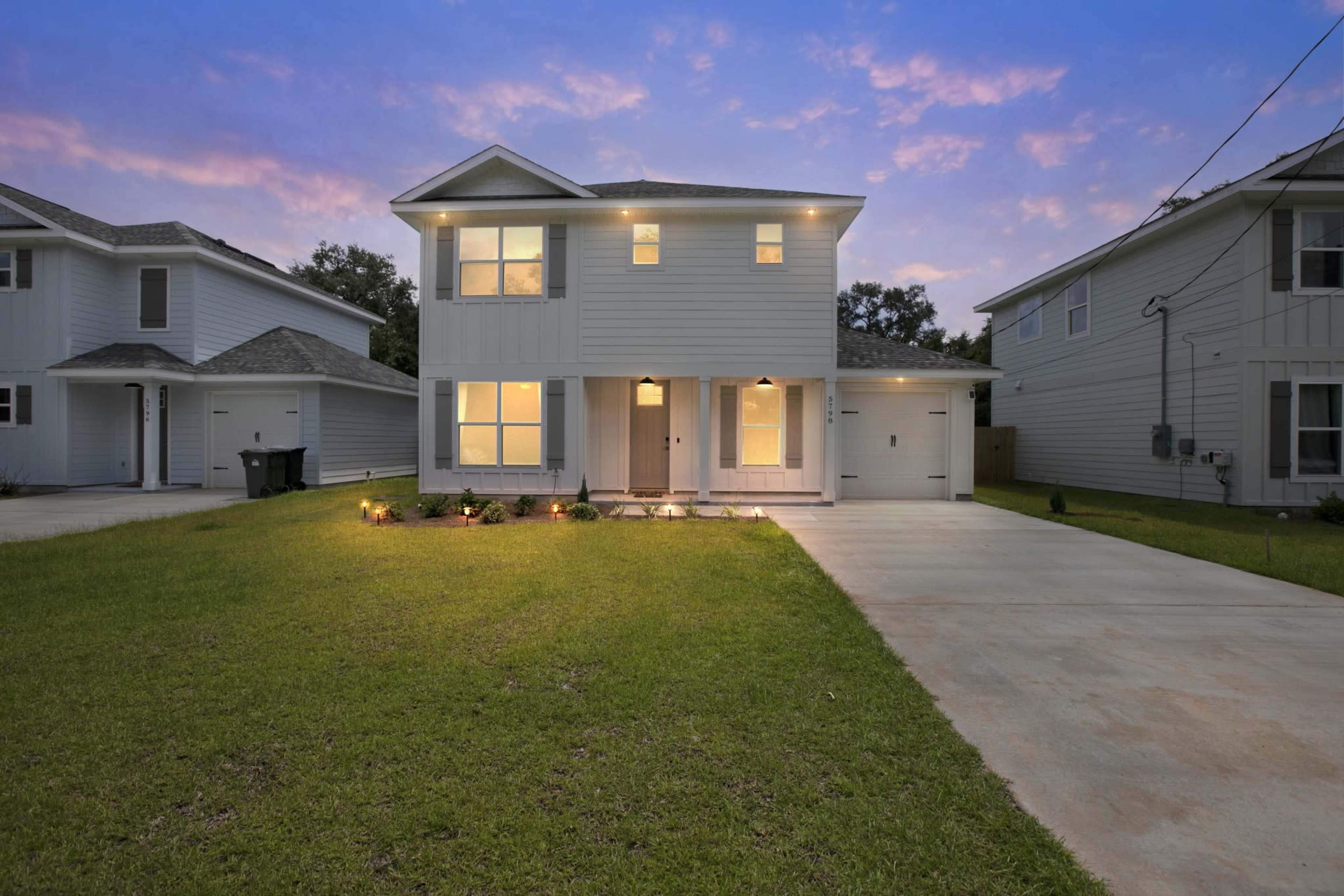 A two-story gray house with a covered front porch, large windows, and a concrete driveway is surrounded by a grassy yard at dusk.