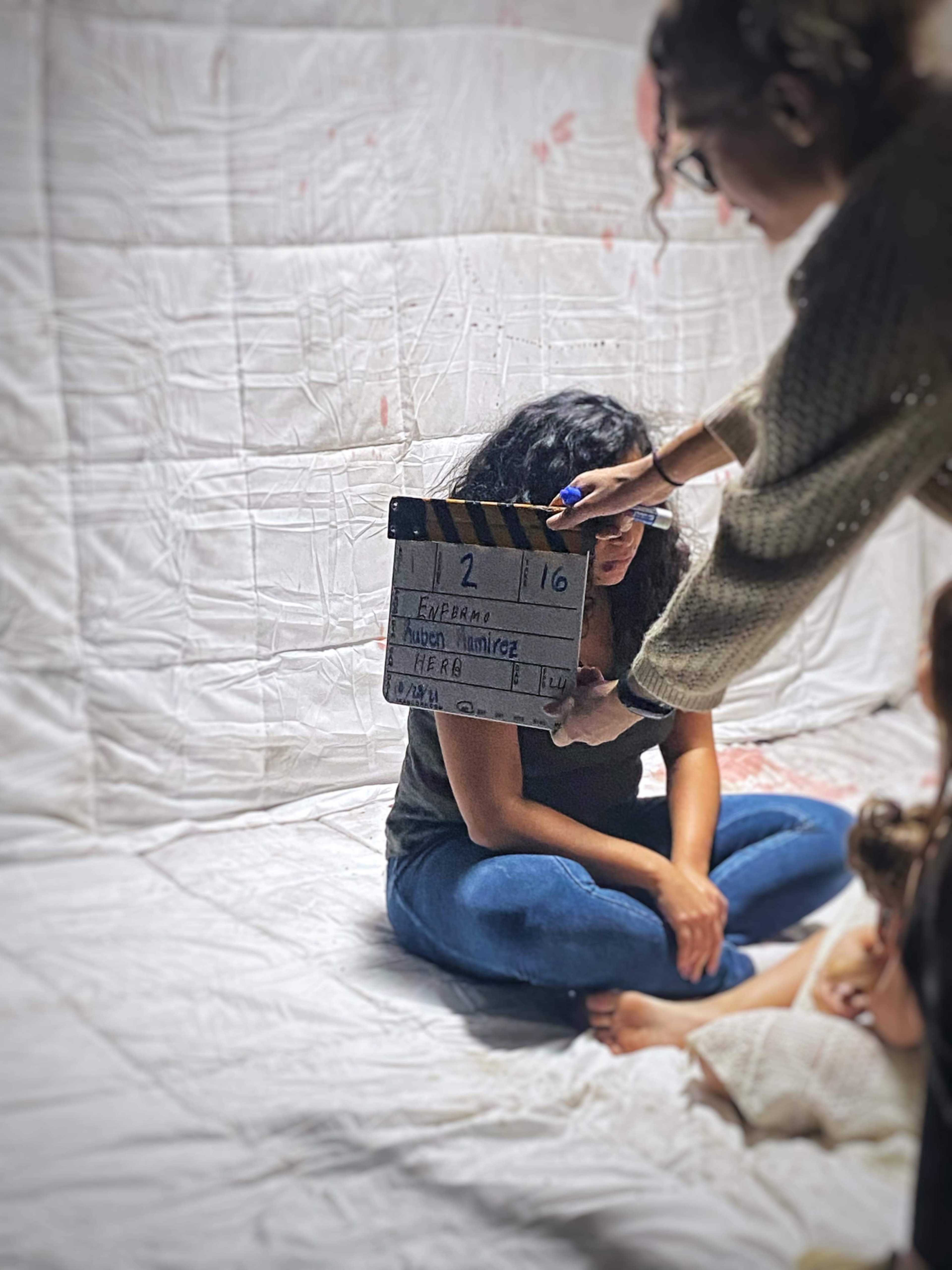 A woman sits on the floor against a white backdrop while another person holds a clapperboard labeled with details for a film scene.