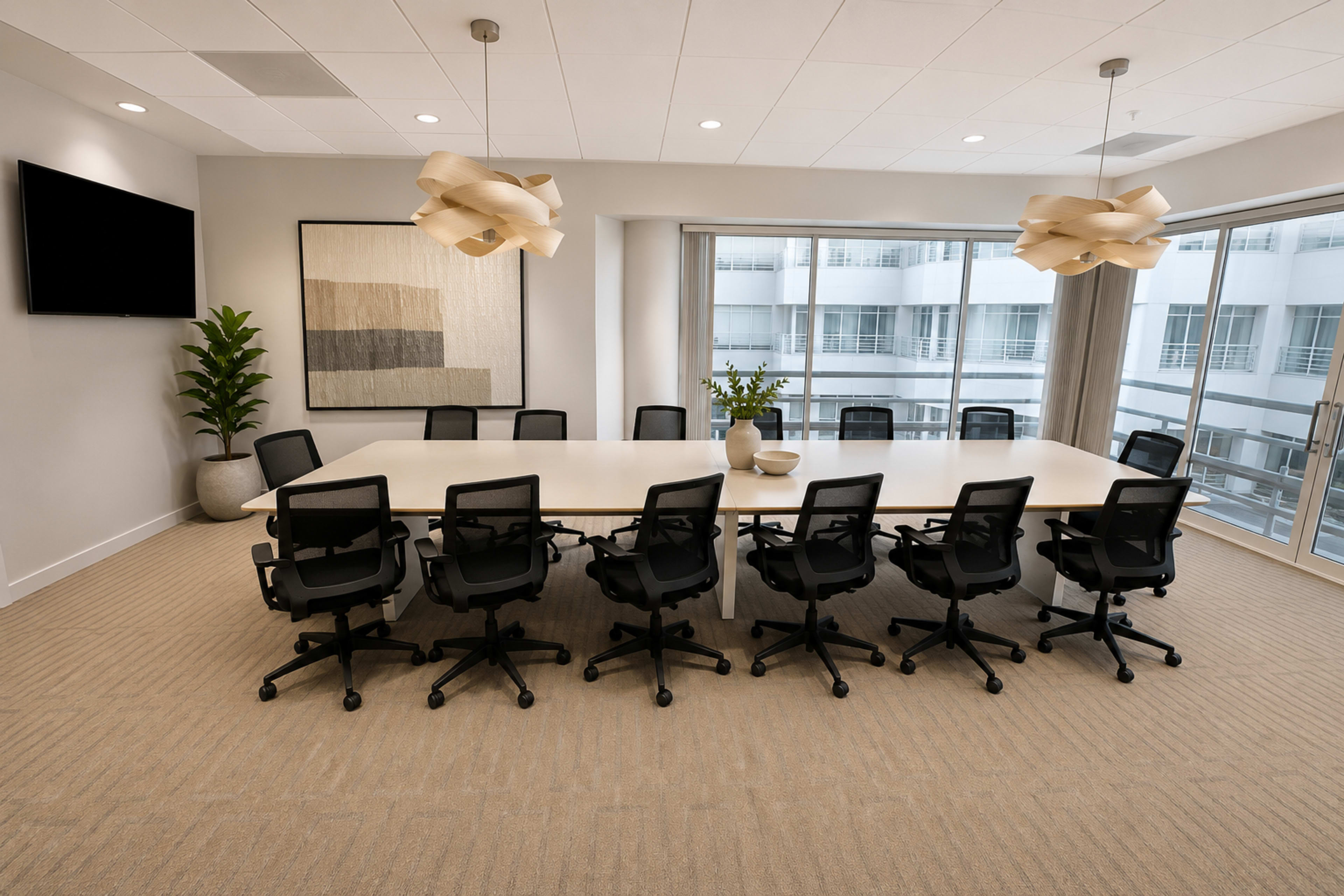 A modern conference room features a large oval table surrounded by black office chairs, with large windows and contemporary light fixtures overhead.