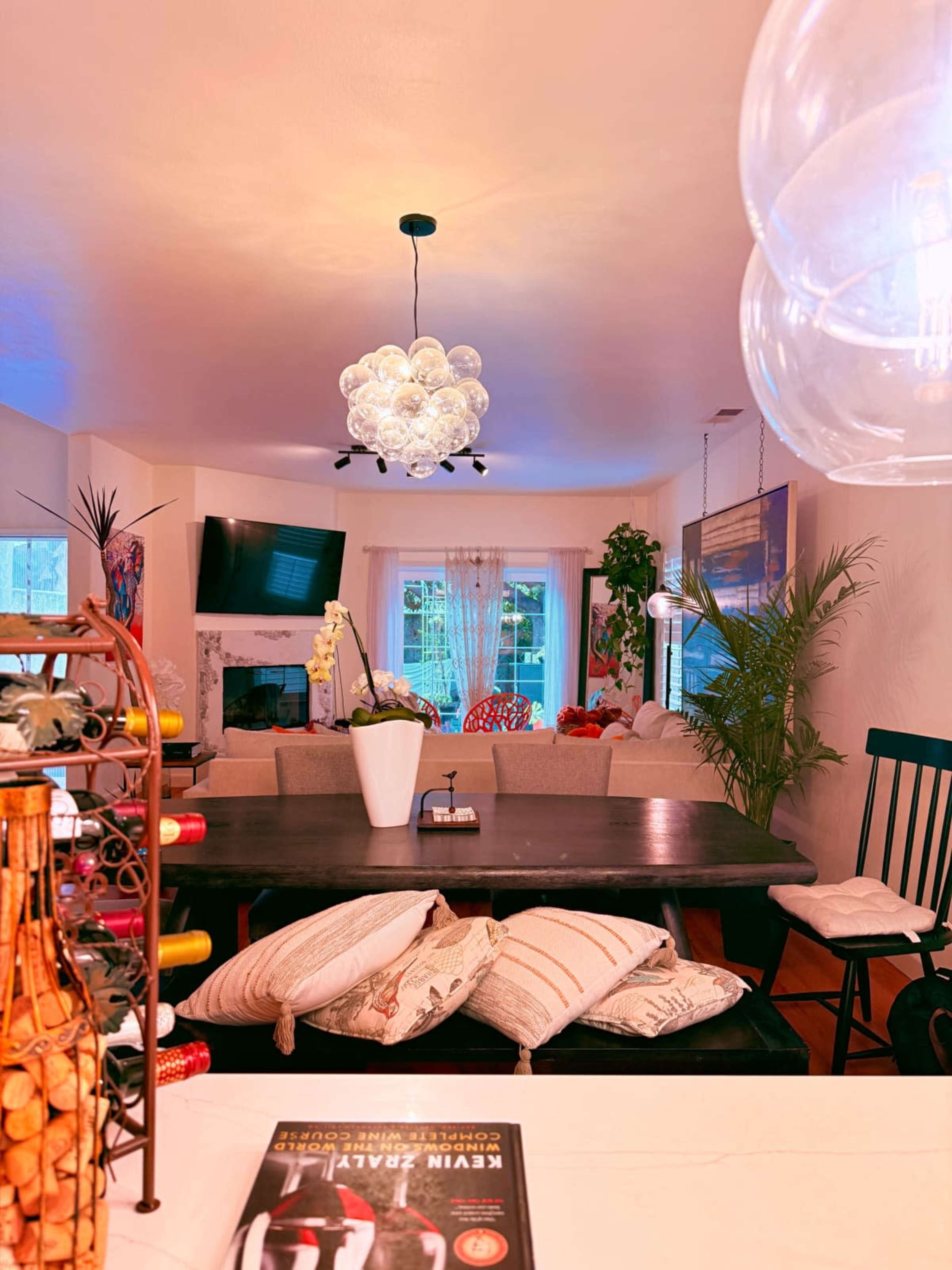The image shows a cozy dining area with a dark wooden table, decorative bottles, and modern lighting, leading into a brightly lit living space with plants and a television.