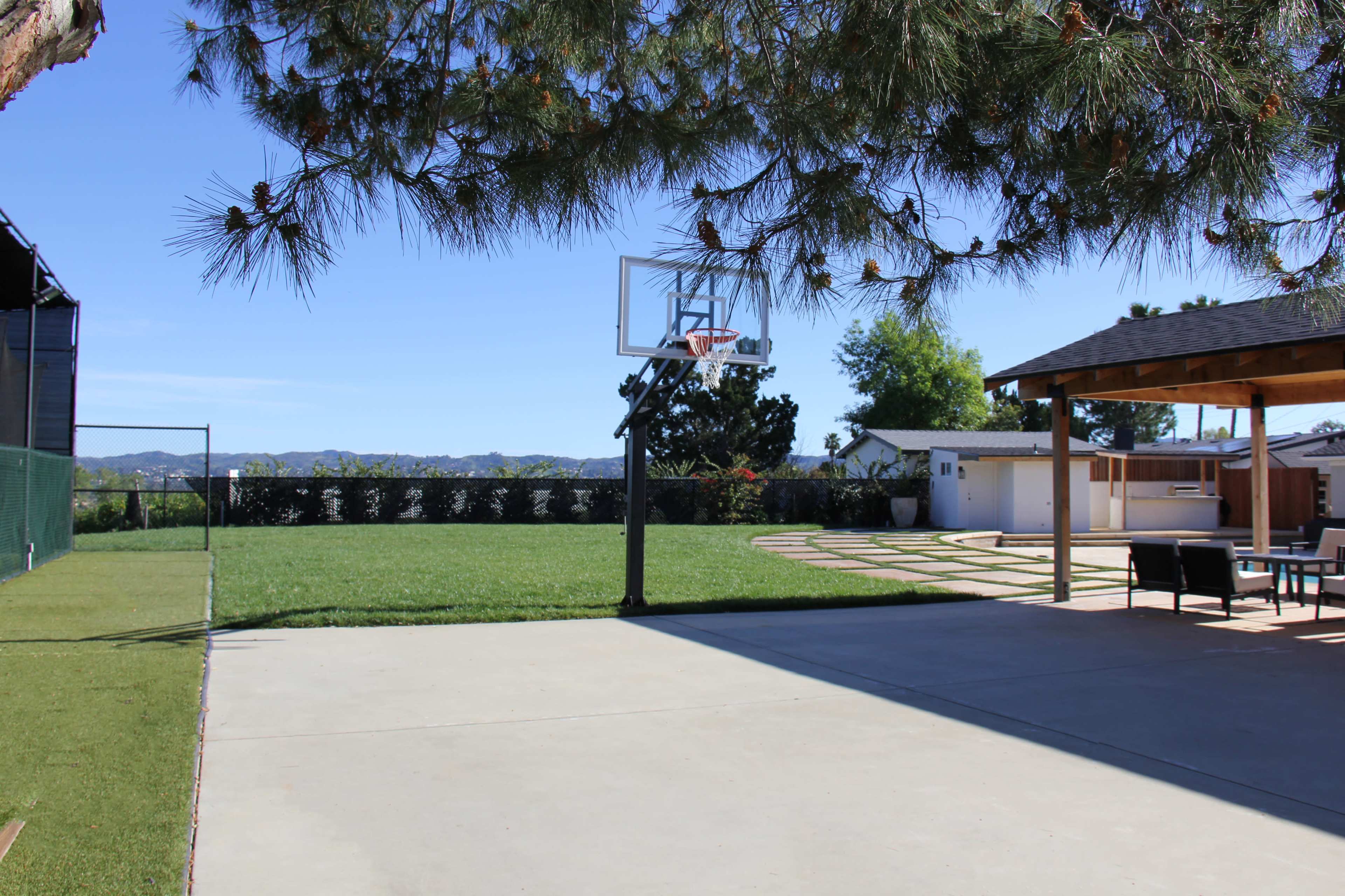 The scene shows a sunny backyard with a basketball hoop, a grassy area, a patio with seating, and a view of surrounding hills.