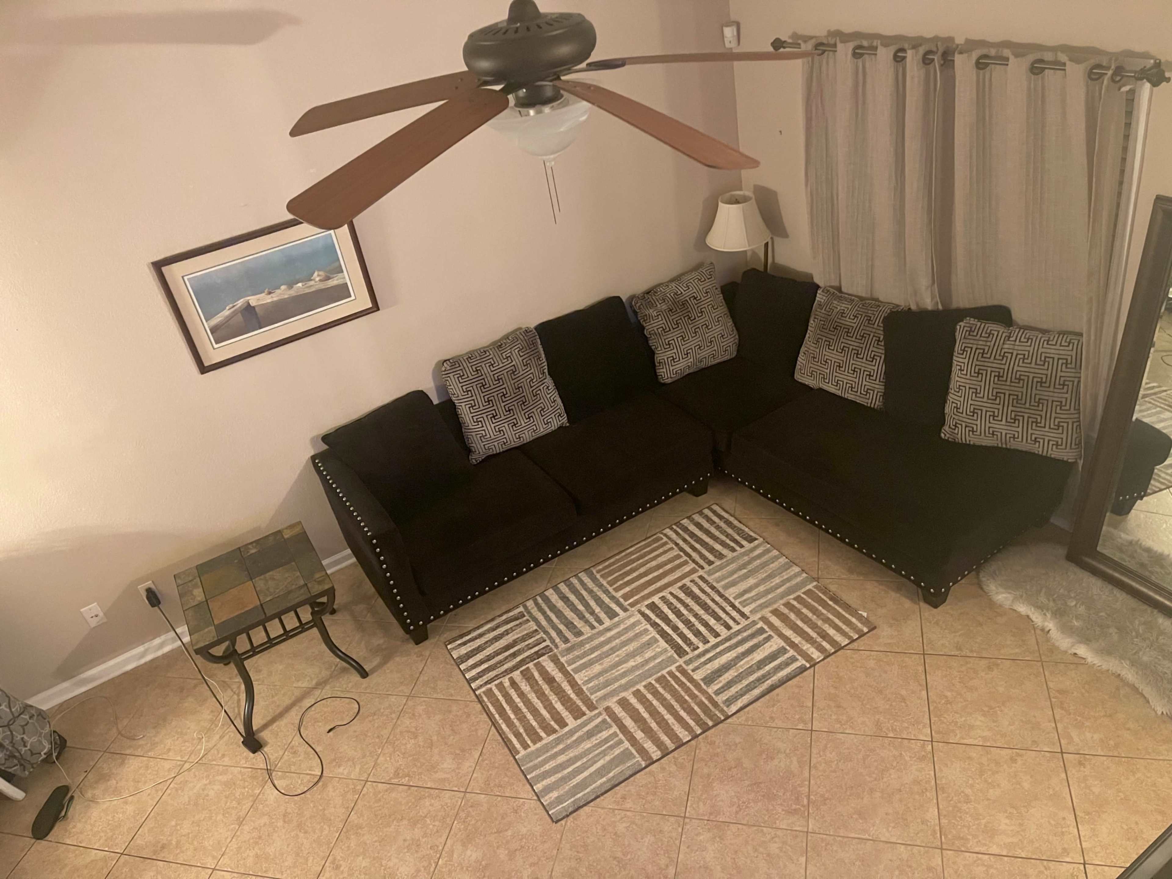A living room with a dark sectional sofa, a patterned area rug, a ceiling fan, and a lamp beside a window.