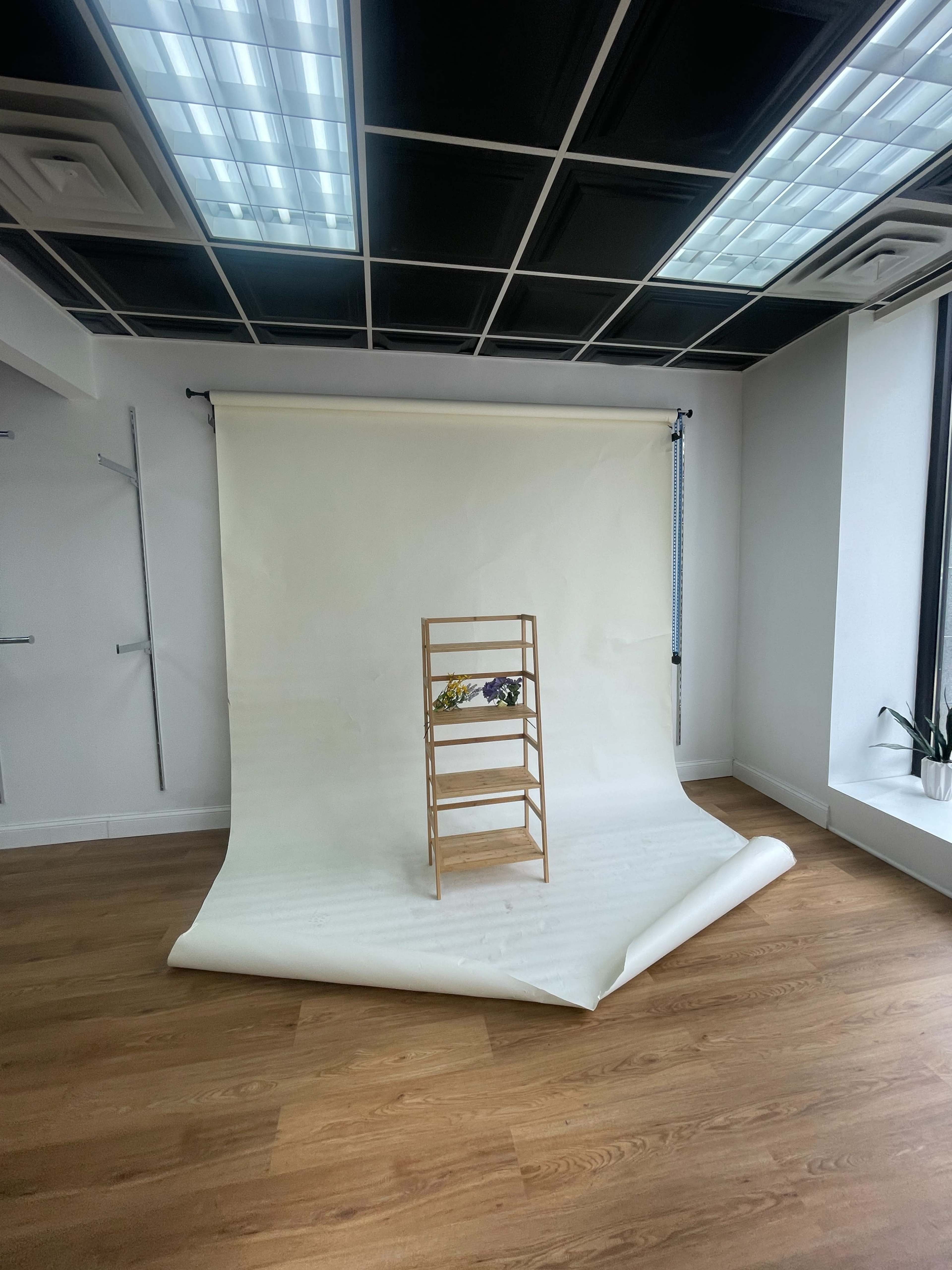 A wooden three-tiered shelf is positioned in front of a white backdrop in a well-lit room with a wooden floor.