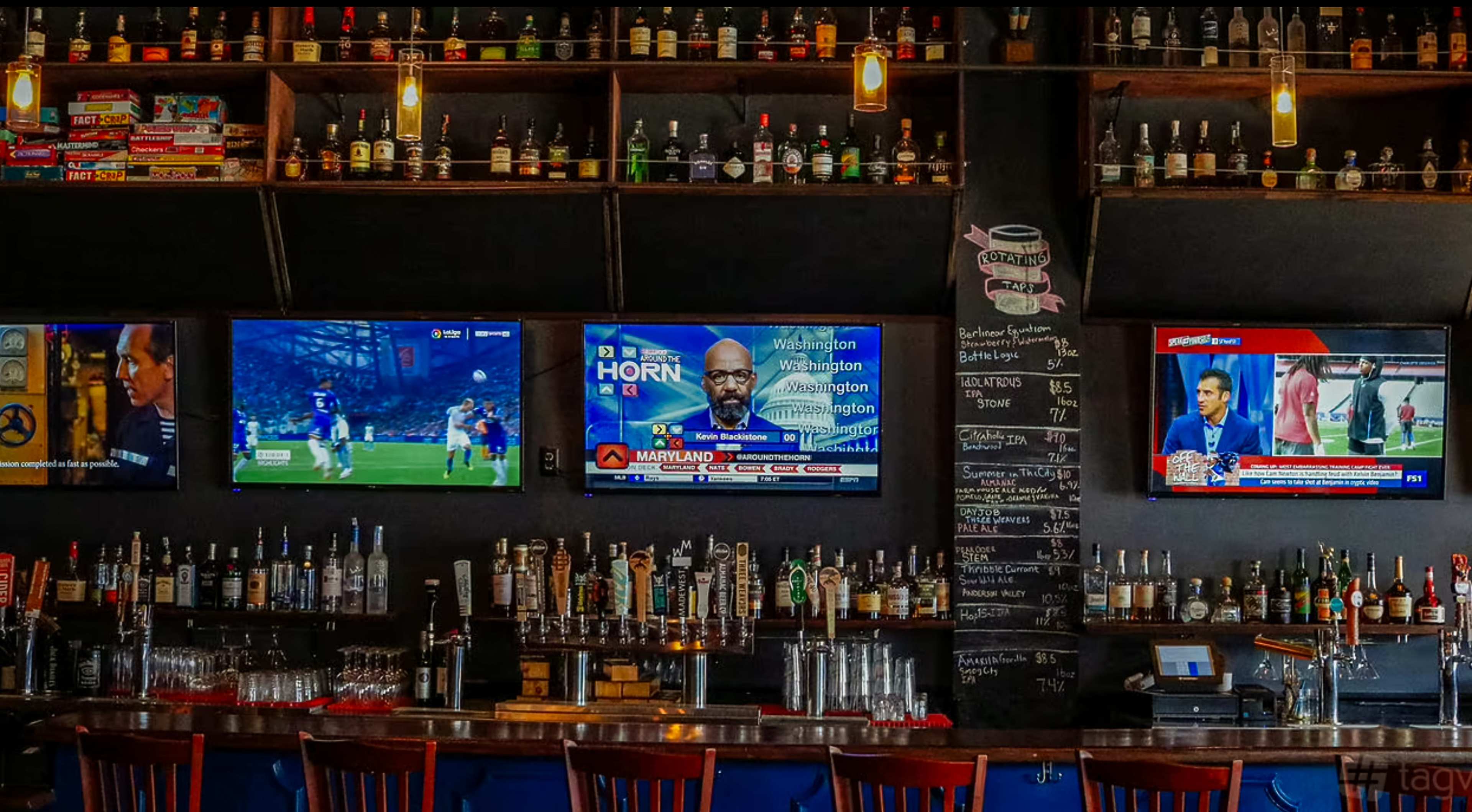 The image shows the interior of a bar with shelves lined with bottles of alcohol, two televisions displaying sports events and news, and a chalkboard menu on one wall.