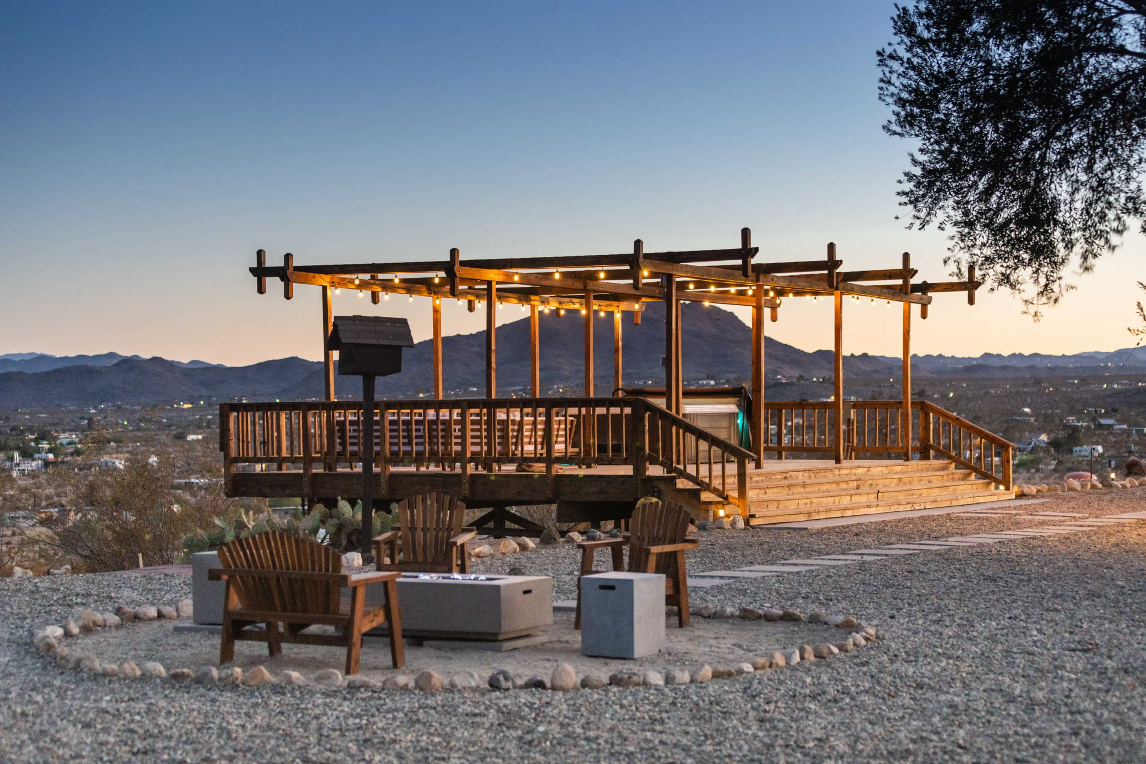 A wooden observation deck with string lights overlooks a valley surrounded by mountains, with stone fire pits and seating arranged at its base.
