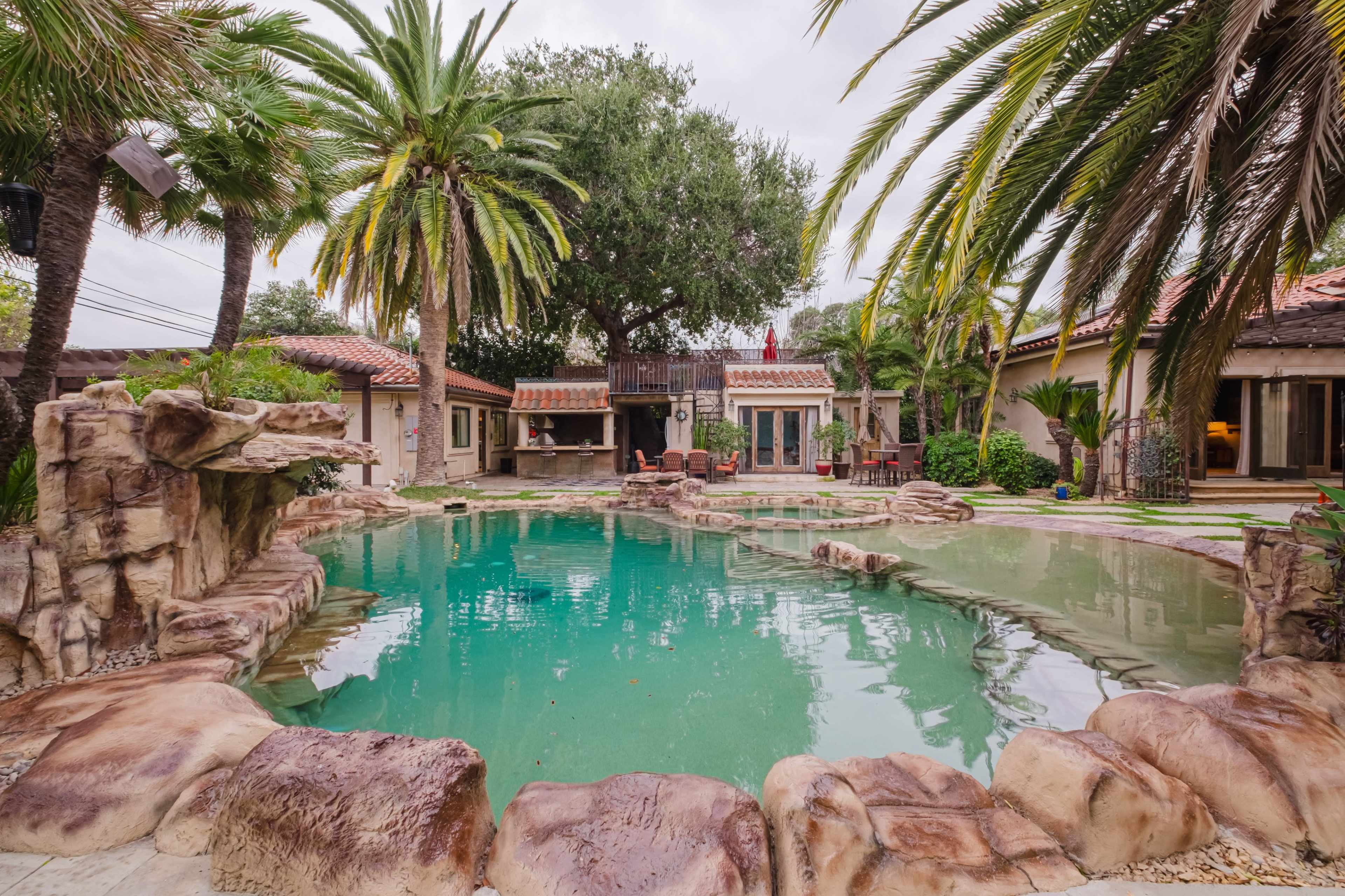 A residential backyard featuring a swimming pool surrounded by palm trees and rock formations, with a house visible in the background.