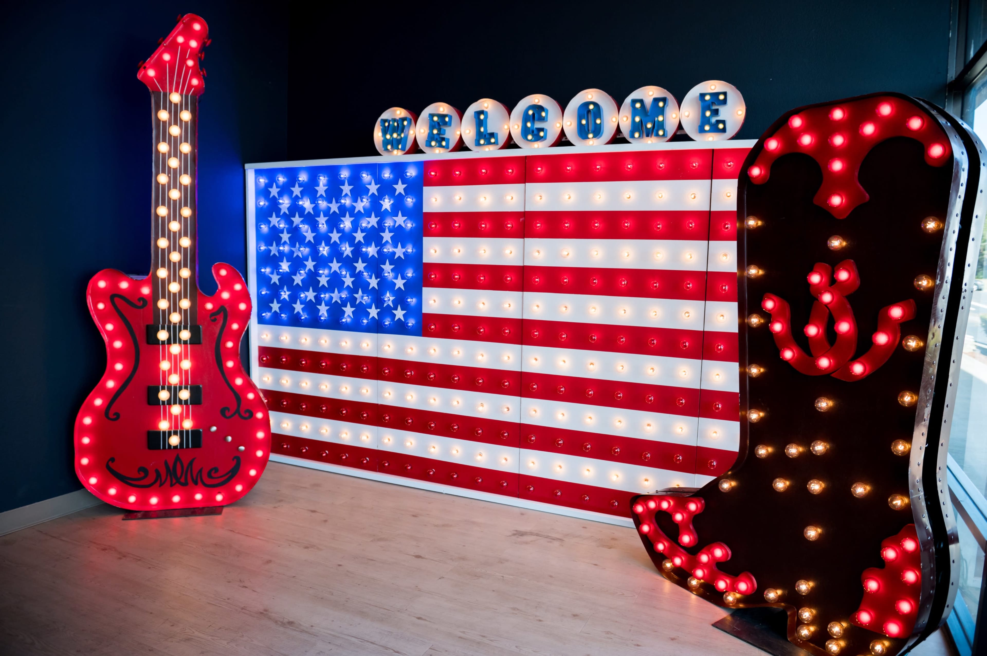 The image features a wall display with a large illuminated American flag alongside a red guitar and a decorative cowboy boot, both adorned with lights, and a "WELCOME" sign above.