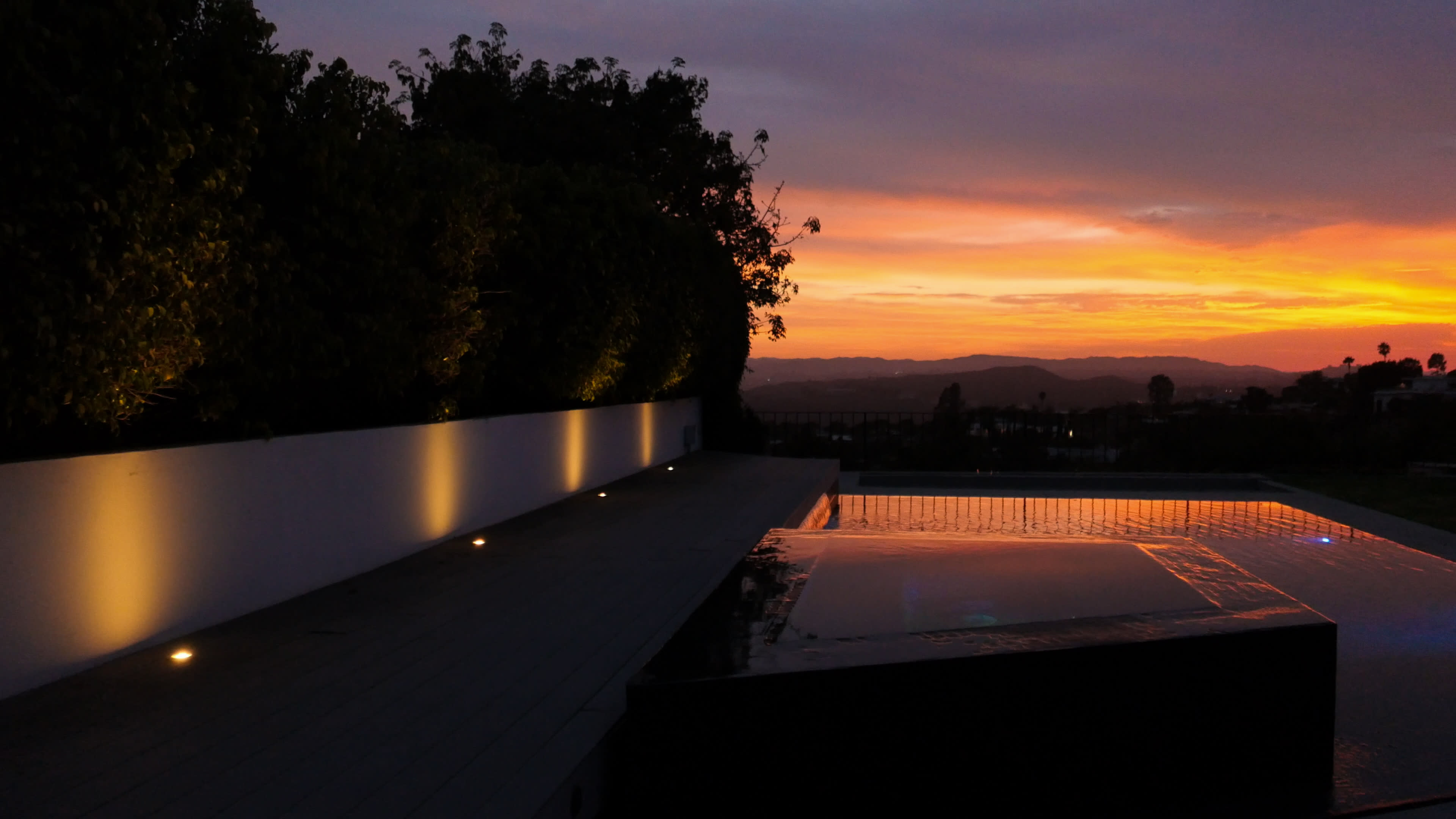 A swimming pool reflects the colorful sunset, surrounded by greenery and softly lit pathways.