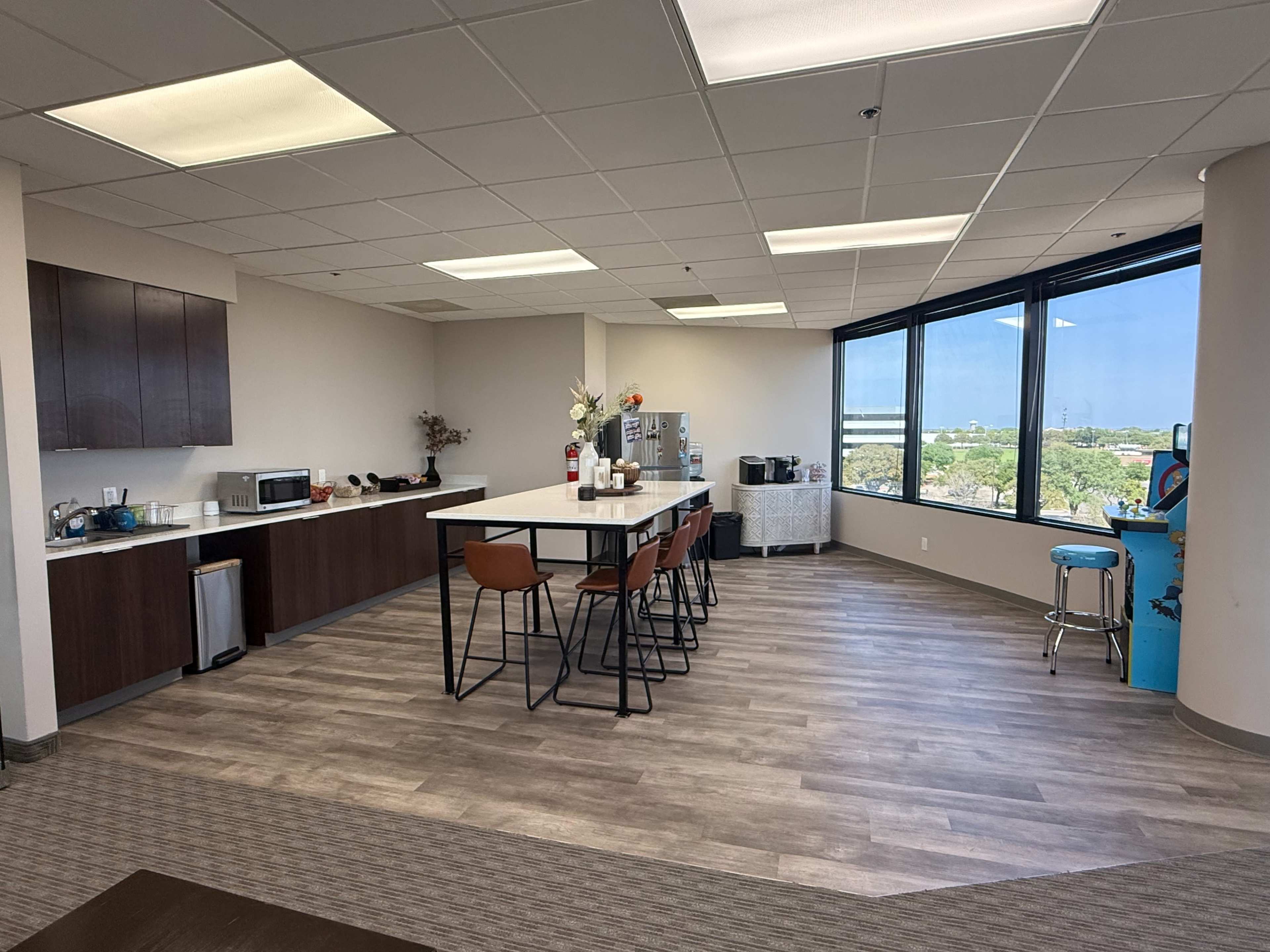 The image shows a modern office break room featuring a kitchen area with a microwave, coffee machine, and seating at a long counter, alongside large windows with a view of the outdoors.