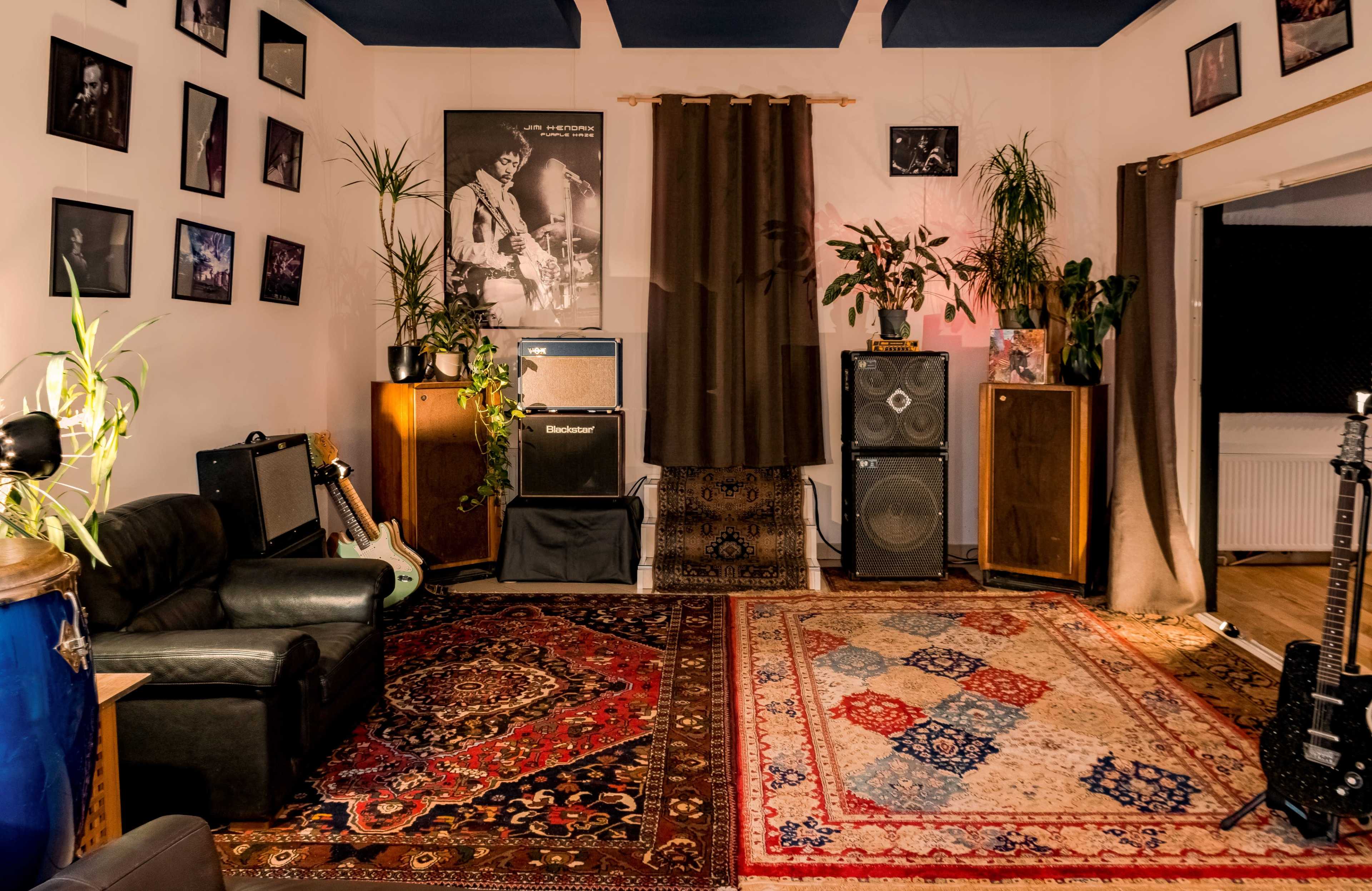 The scene shows a music room featuring a black sofa, guitars, large speakers, a patterned rug, and various framed photos on the walls, all surrounded by indoor plants.