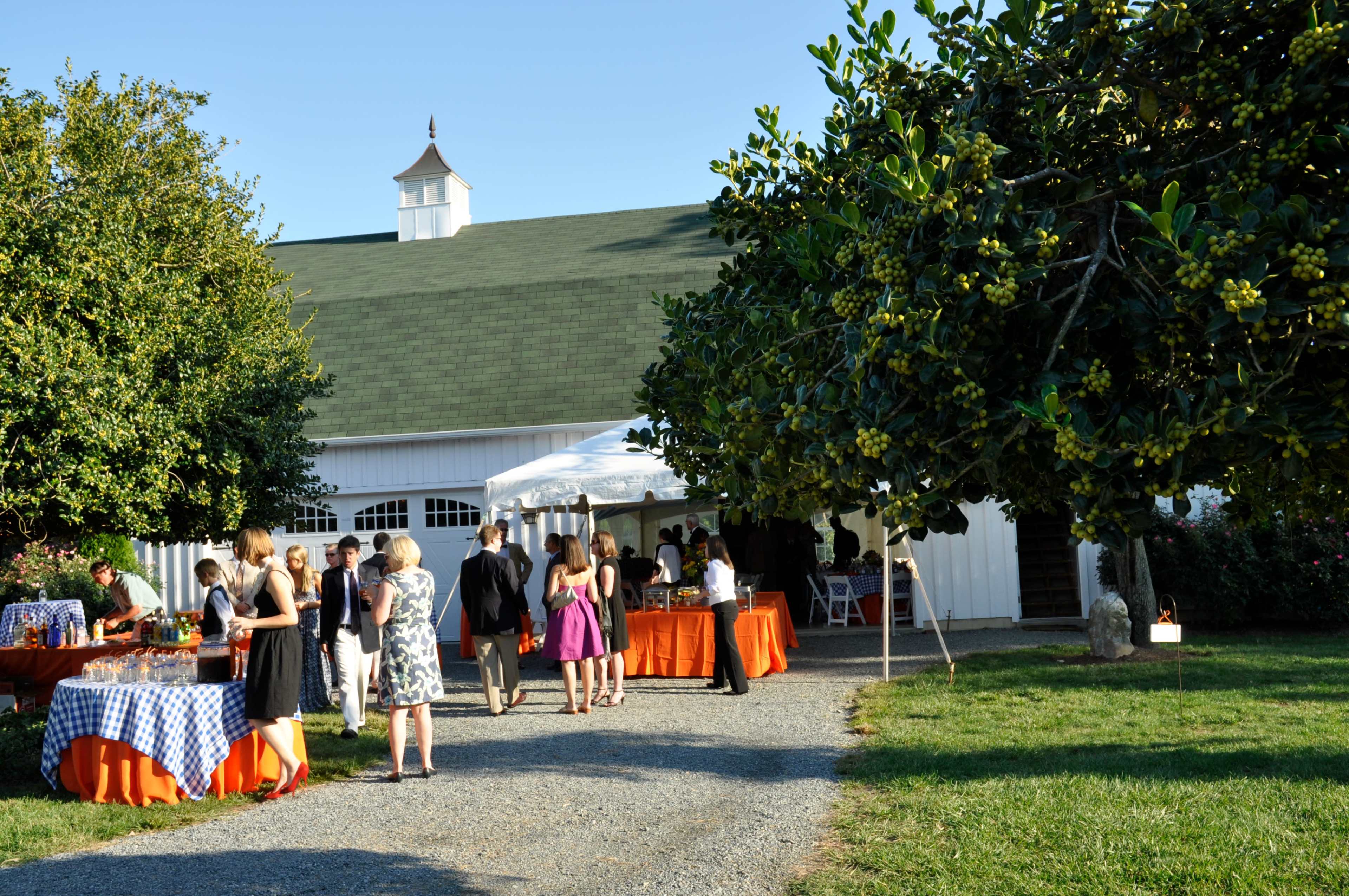 A group of people mingles outside a barn-style building during a social event, with tables set under a tent and a tree in the foreground.