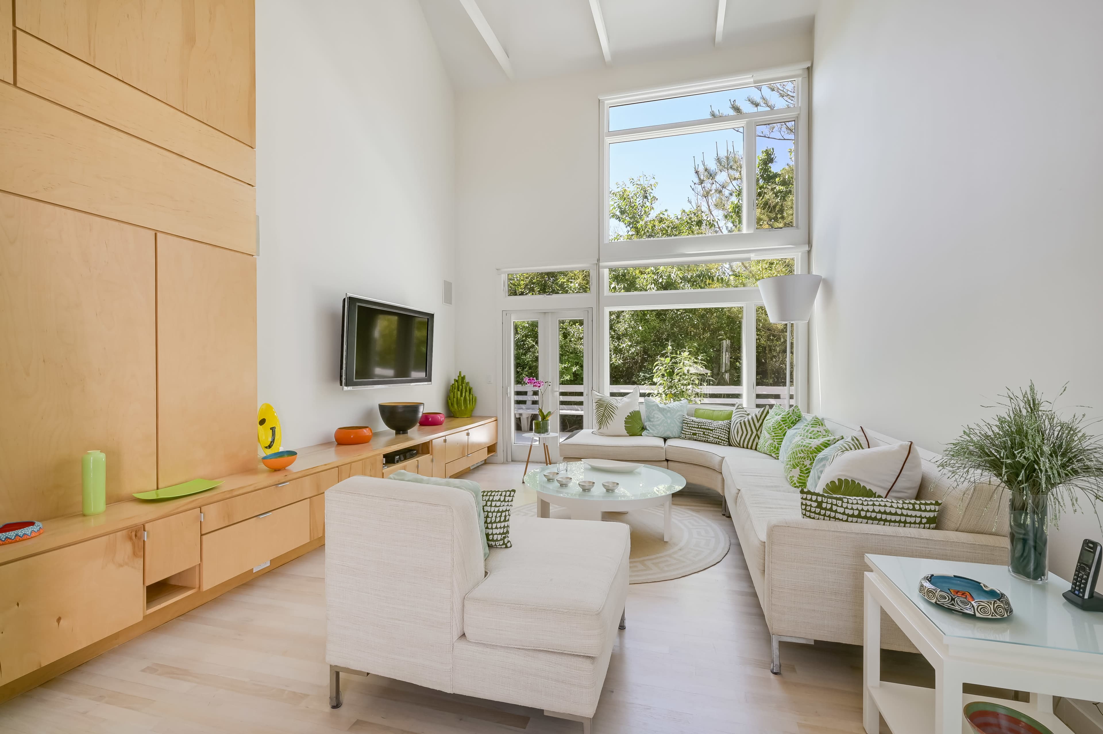 A bright living room features a sectional sofa, a television mounted on the wall, and large windows overlooking greenery.